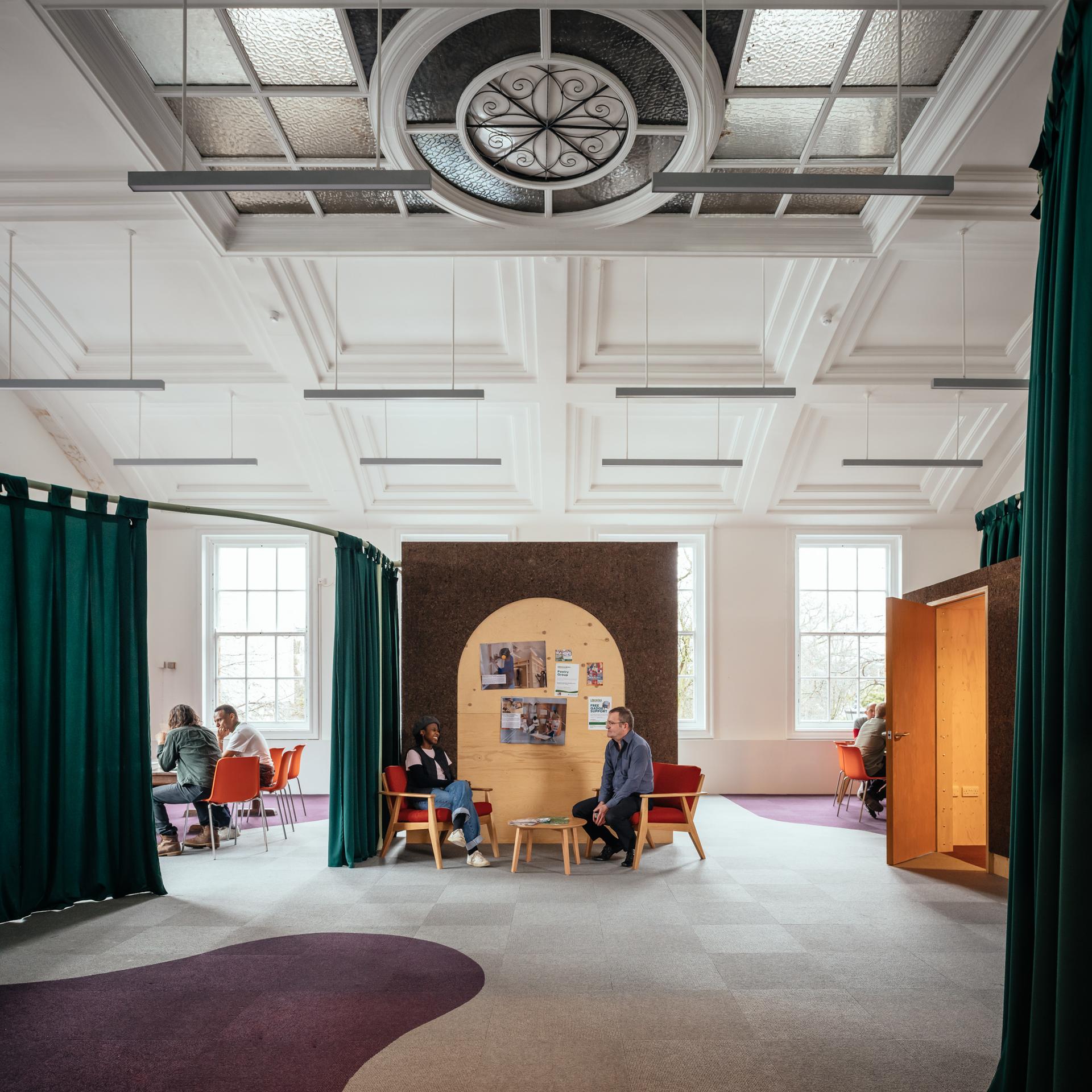 A spacious, modern room with high ceilings and large windows. Spruce plywood accents add warmth as people talk in groups; two sit under a circular noticeboard, others gather at tables. Green curtains frame the timber construction.
