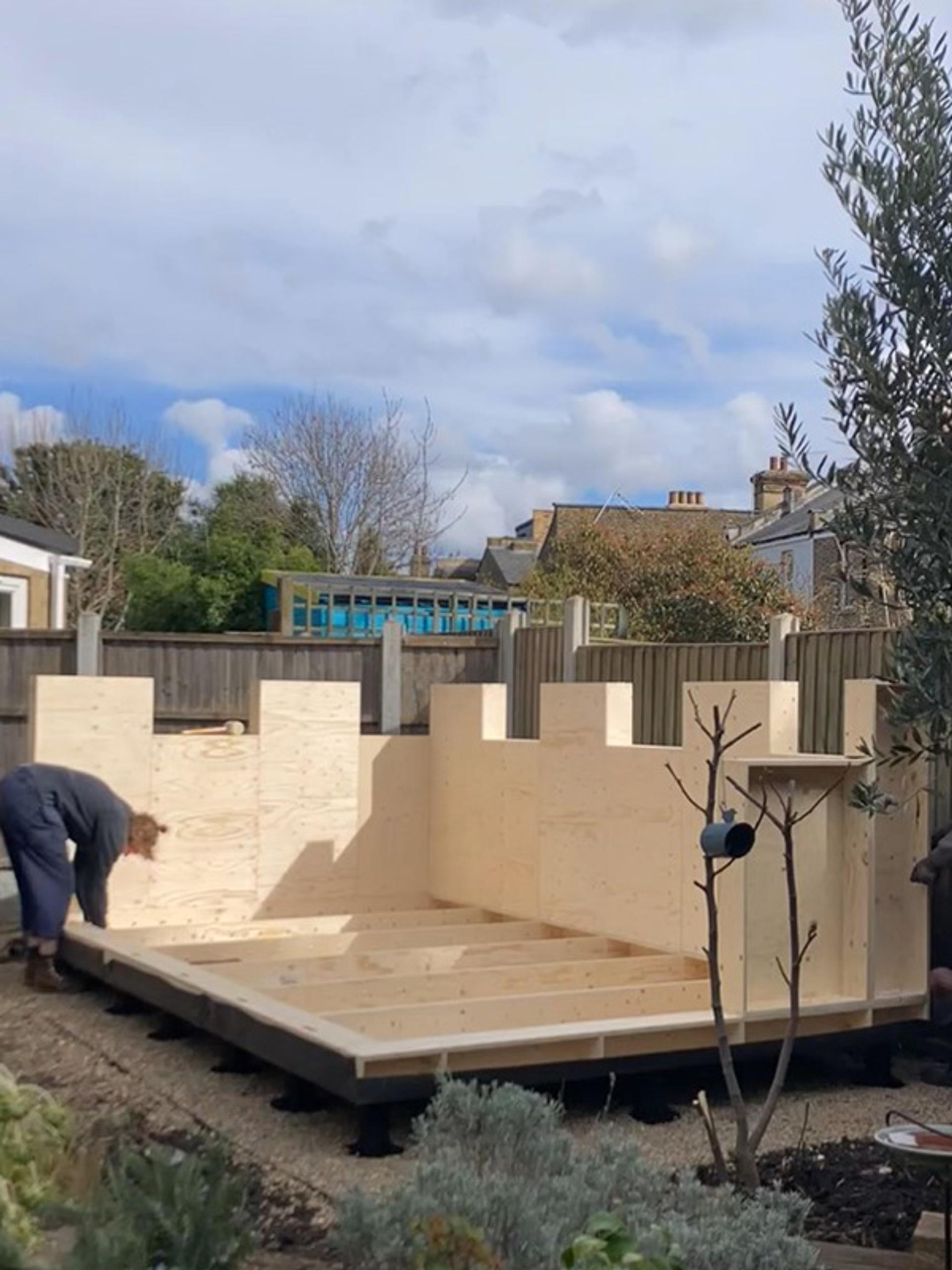 A person is bent over, working on assembling the sustainable spruce walls of a wooden structure in a garden. The unfinished building has notched tops, with surrounding houses and trees visible under a partly cloudy sky.