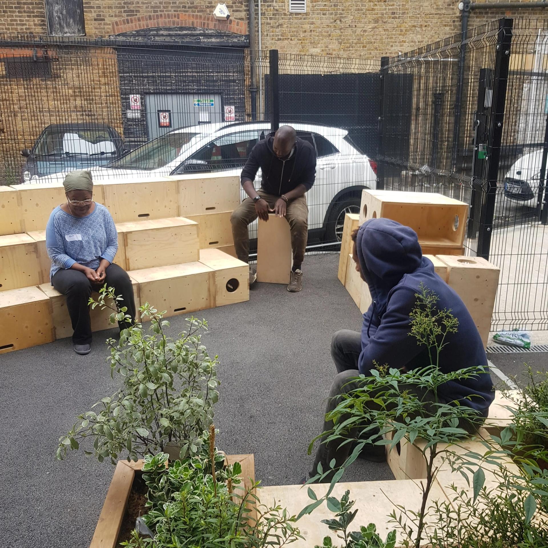 Three people sit apart on wooden benches made from recycled materials in an outdoor urban area surrounded by plants, with a brick building and a parked white car in the background. The quiet atmosphere reflects values of reuse and mindful living.