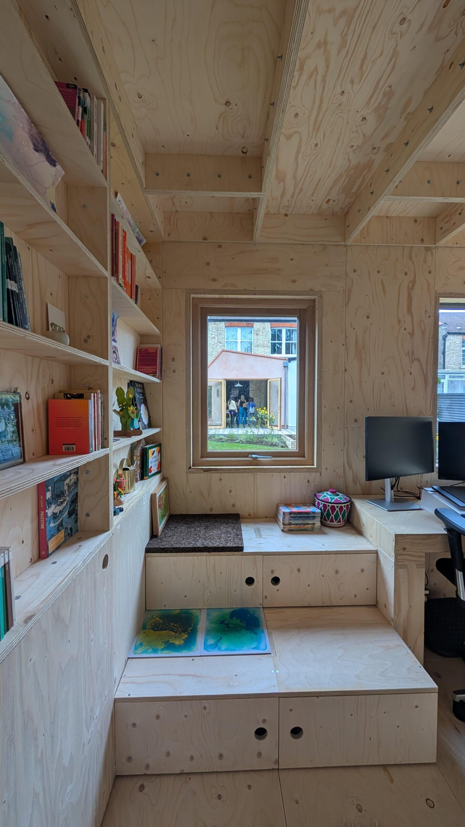 A cosy, light wood-panelled U-Build home office with built-in shelves, books, and steps leading to a window. Two people are visible outside through the window. A computer, chair, and colourful basket are on the right-hand desk.
