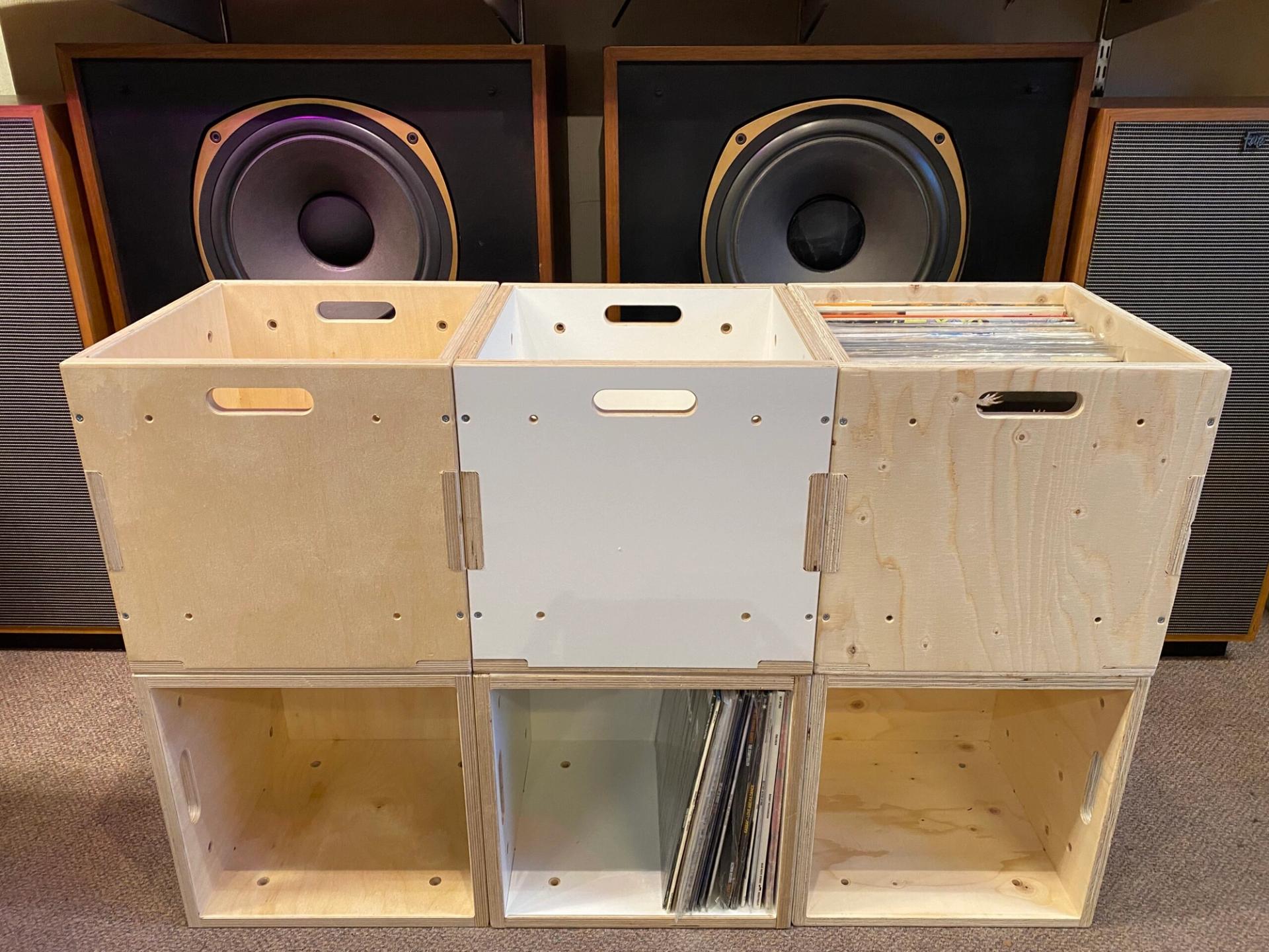Six wooden crates, some natural and some painted white, are stacked in two rows. Several crates contain vinyl records. Made from sustainably sourced wood, they sit in front of two large loudspeakers with visible cones in the background.