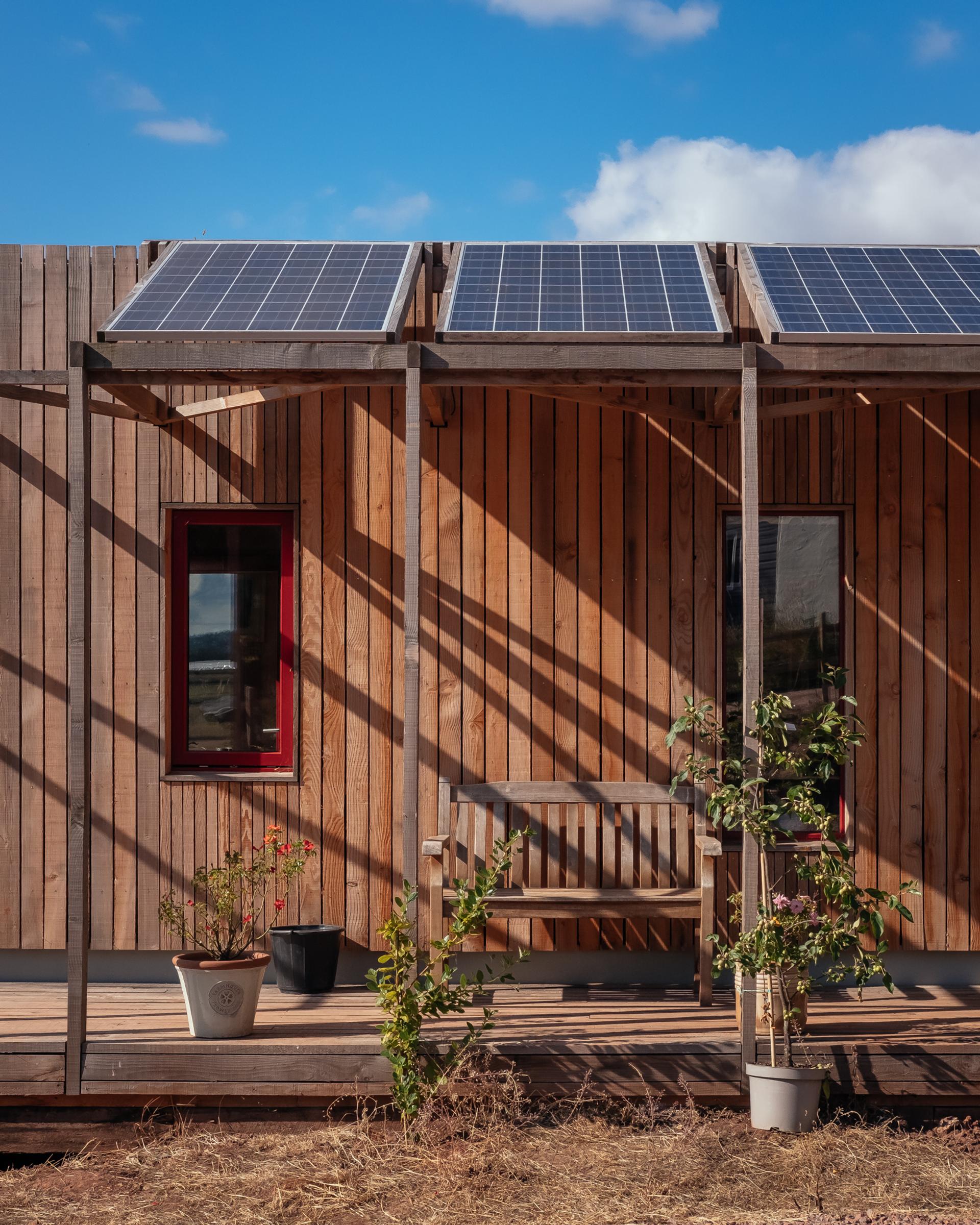 A small modular wooden house made of birch, with solar panels on the roof, a red-framed window, and a porch with a bench and potted plants under a blue sky.