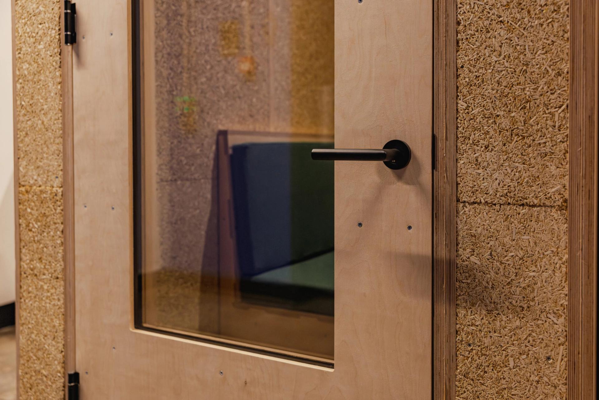 A close-up of a U-Build wooden door with a glass window and black handle, revealing part of a furnished room with a blue and green seat inside. The door is set in a textured wall.