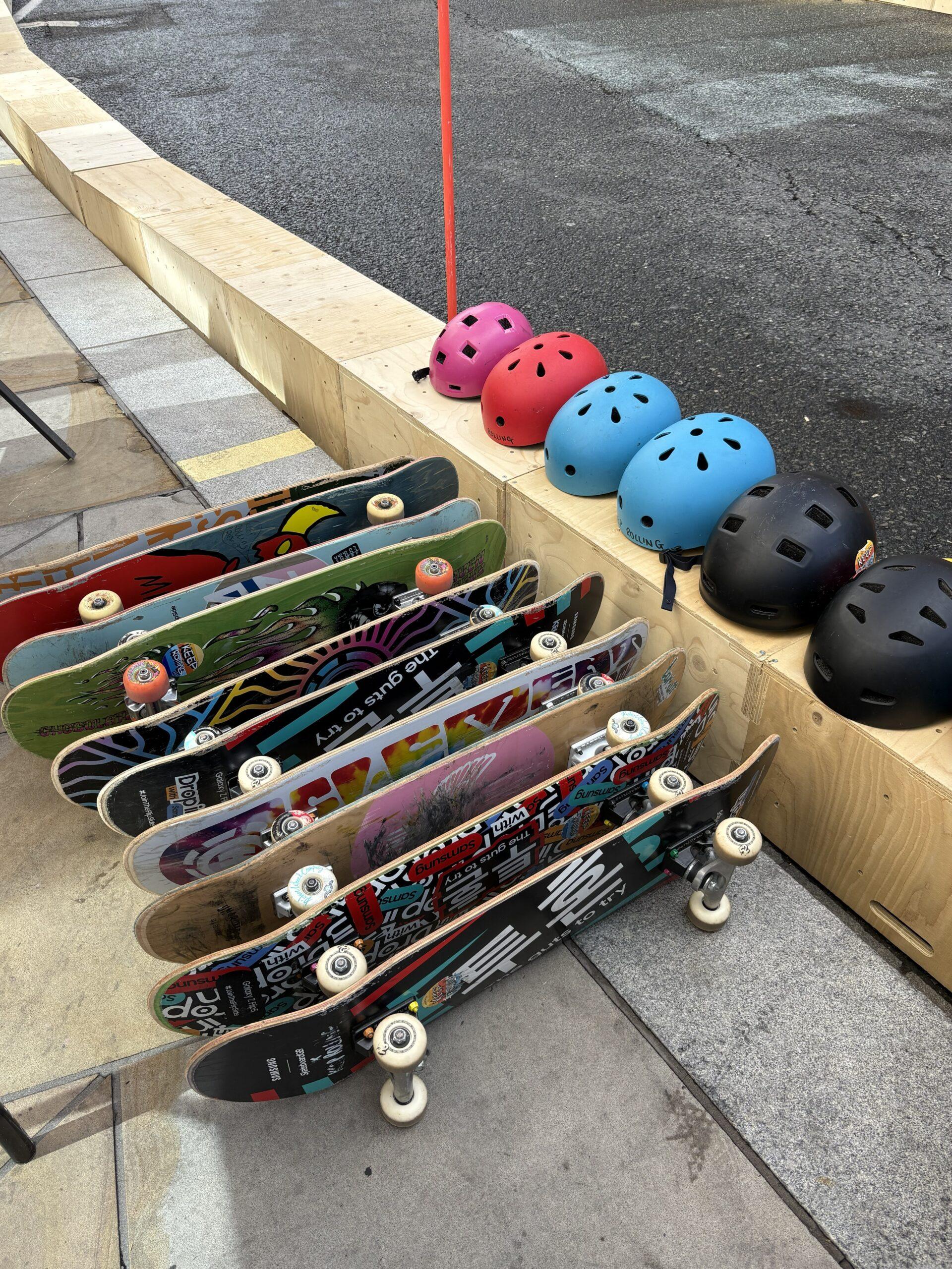 Several colourful U-Build skateboards are stacked upright next to a wooden barrier, with six helmets—pink, blue, and black—lined up on top. The scene is outdoors on a paved street.