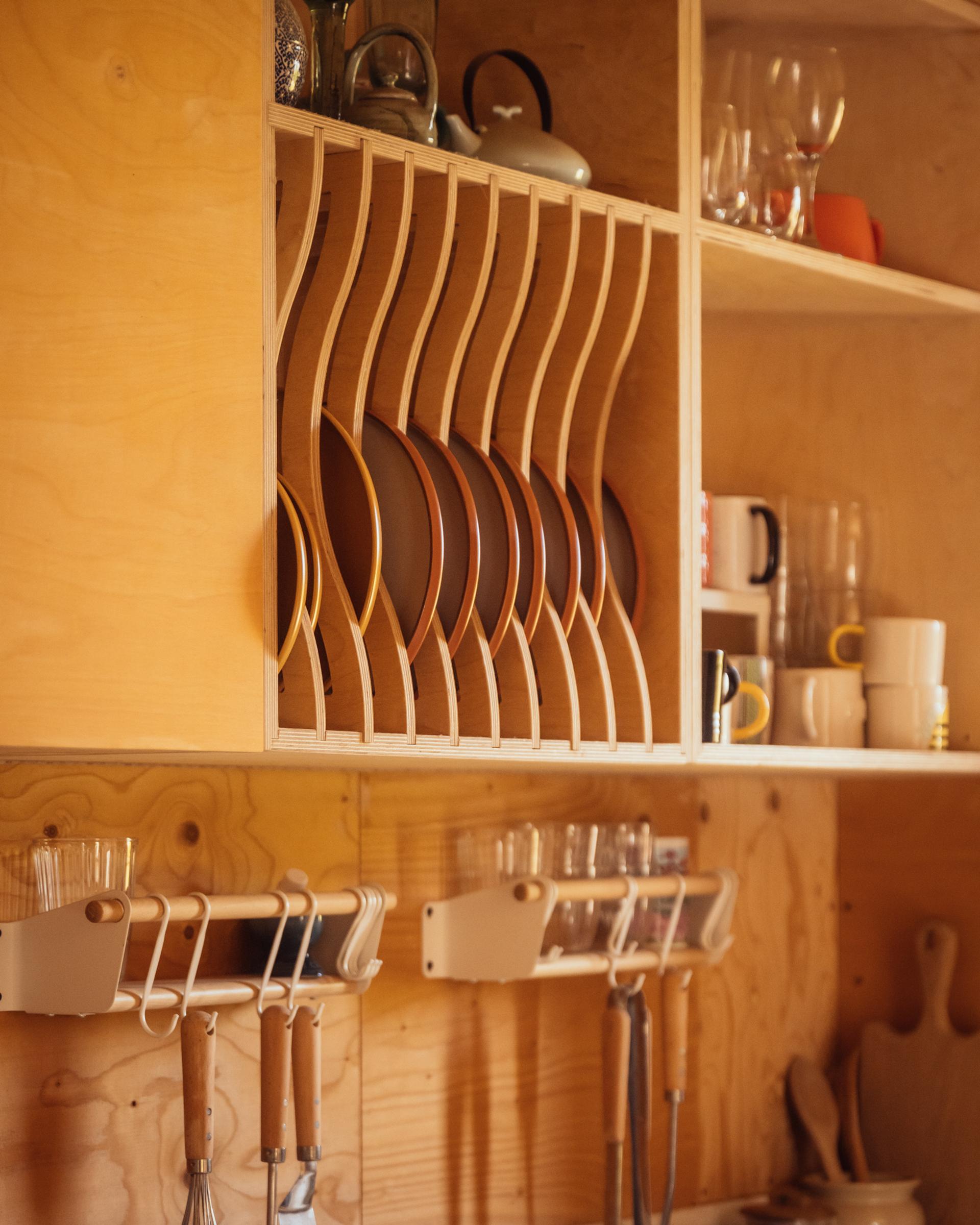 Plywood kitchen cabinetry in a box house extension features a built-in vertical plate rack, with assorted glassware on adjacent open shelves and hanging utensil racks below.