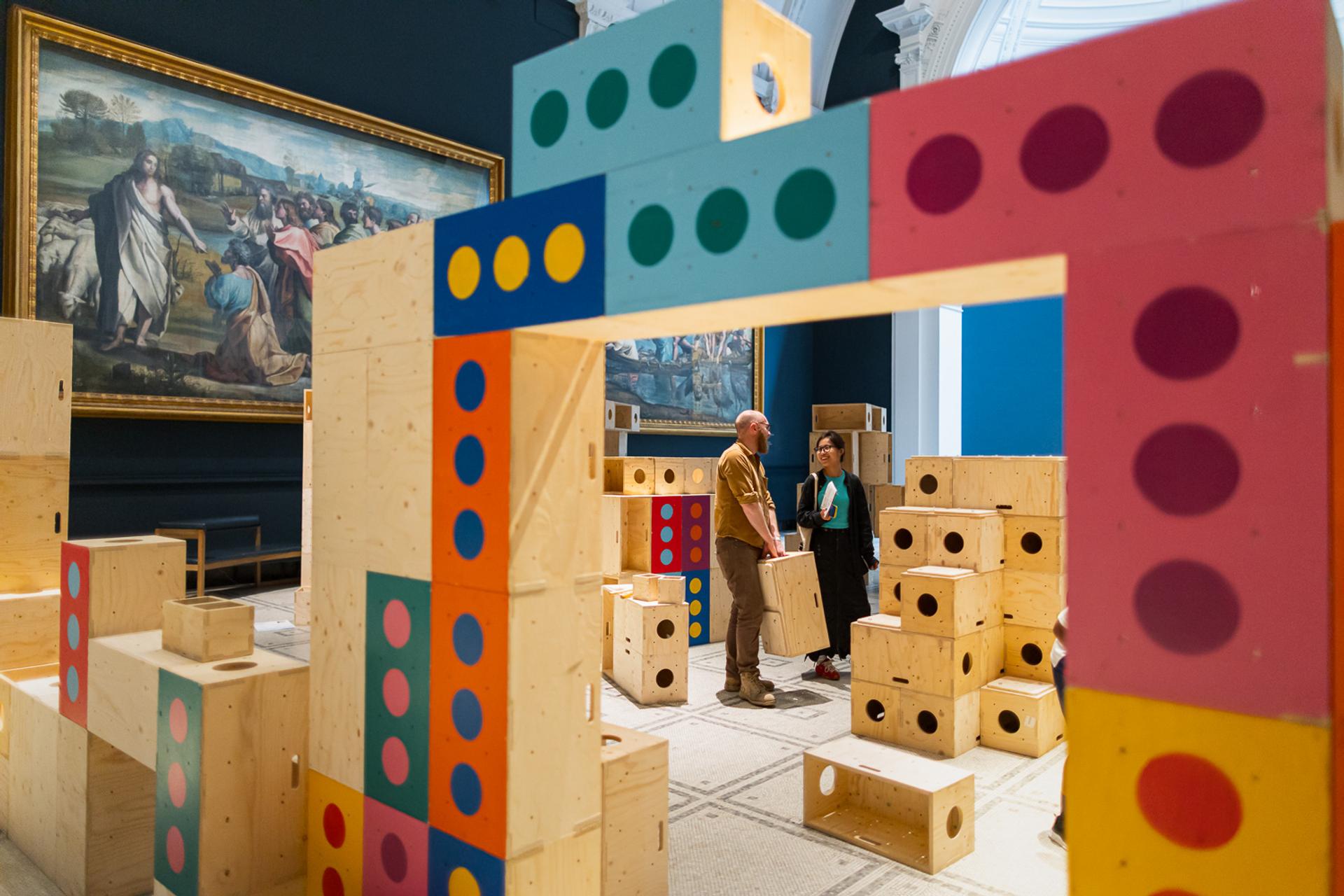 Two people stand among large, colourful wooden blocks in a gallery space at the V&A, surrounded by classical paintings on the walls. Engaging with the interactive Upstart exhibit, they collaborate to build structures for activism, creating a dynamic dialogue between art and action.