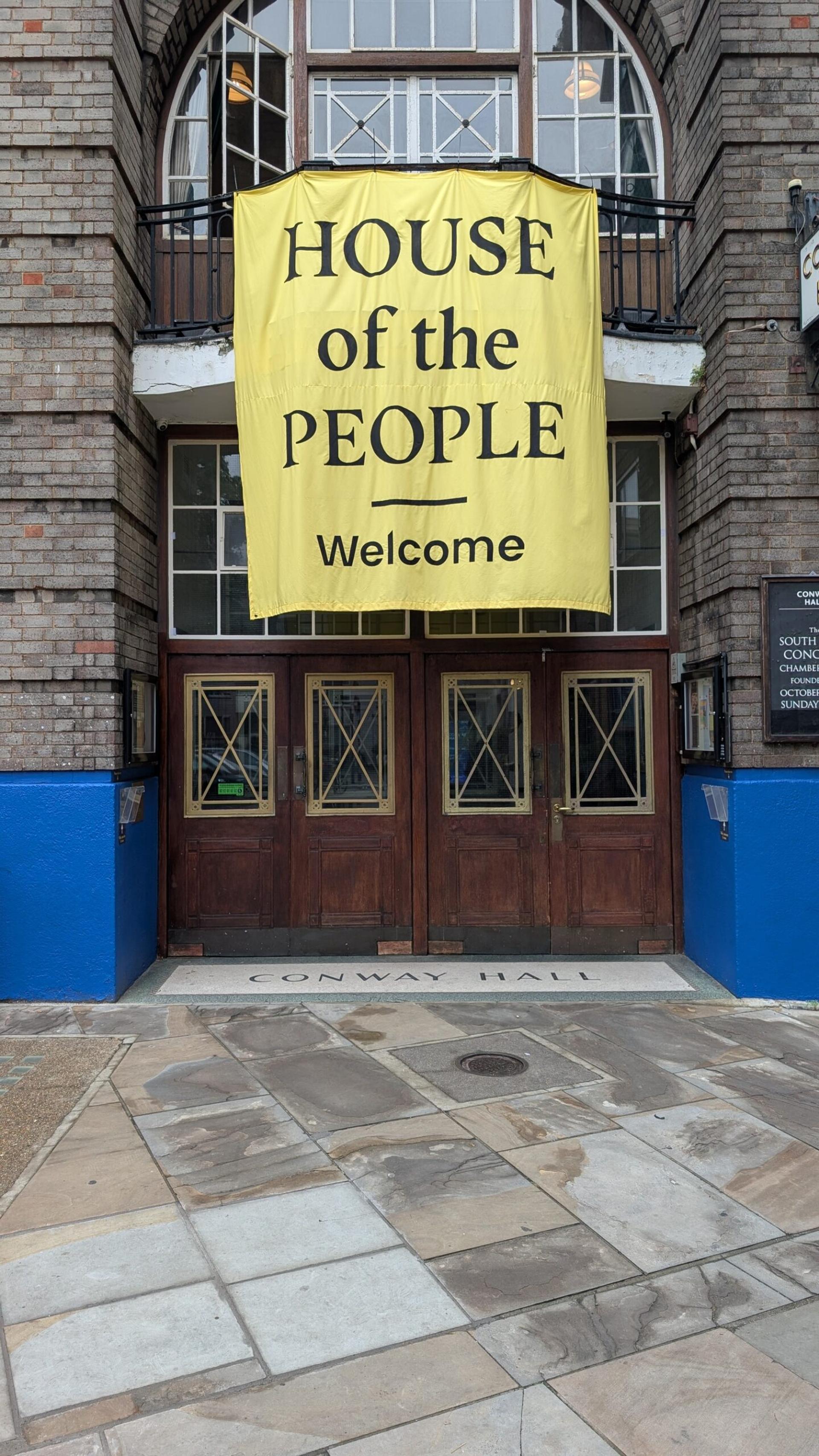 A large yellow banner reading HOUSE of the PEOPLE Welcome hangs above the entrance of a brick building with wooden doors and blue accents. The pavement in front, sustainably sourced for a circular economy, reads CONWAY HALL.