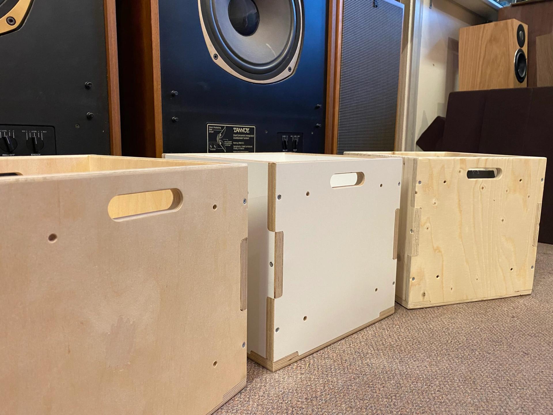 Three wooden storage boxes with handles, one featuring a white panel, are set on a carpeted floor before large black speakers and audio equipment—perfect for anyone embracing the circular economy at home.