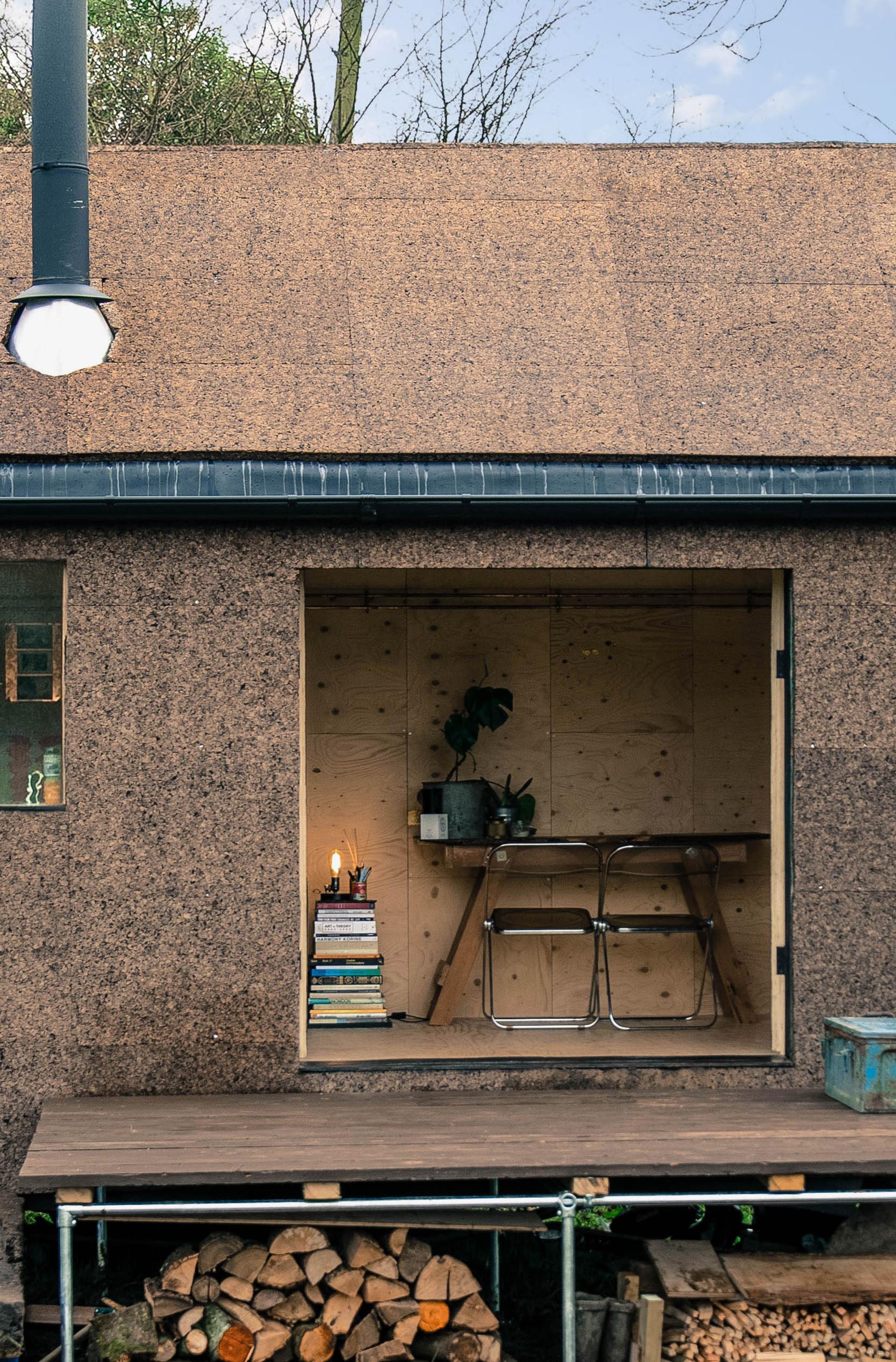 A cosy wooden workspace with a desk, chair, potted plant, stack of books, and lit candles—crafted from sustainably sourced materials—seen through a large square window of a small cabin with a sloped roof and woodpile beneath the porch.