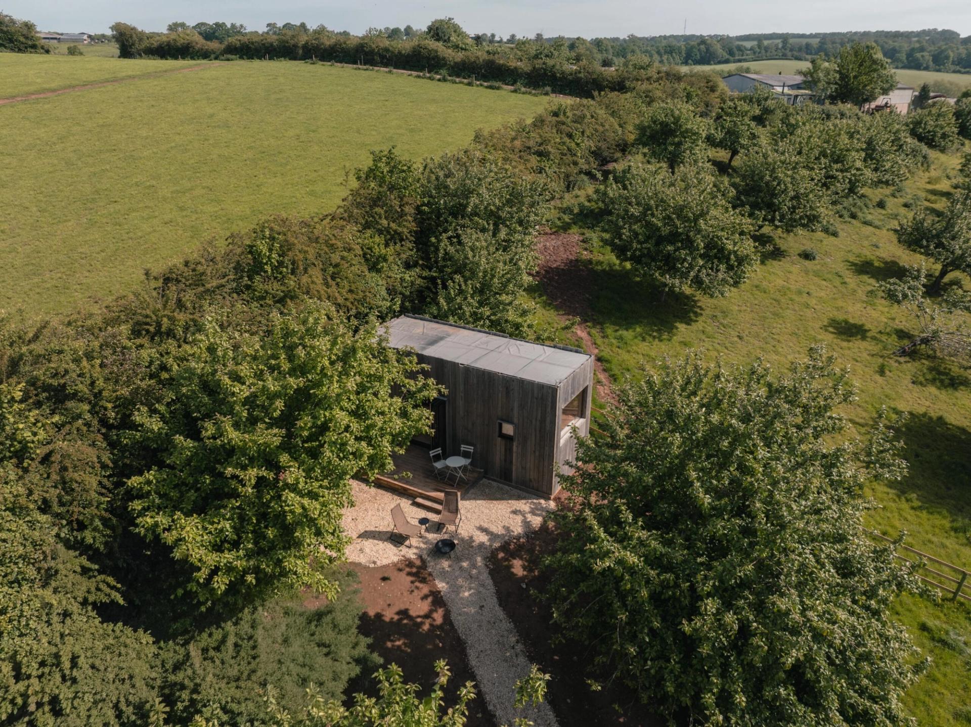 A small, modern wooden cabin sits among green trees and grassy hills, viewed from above on a sunny day. Outfitted with stylish cabin furniture on the veranda, the space invites relaxation. A gravel path leads to the cabin, with outdoor seating nearby. Fields and hedges stretch into the distance.
