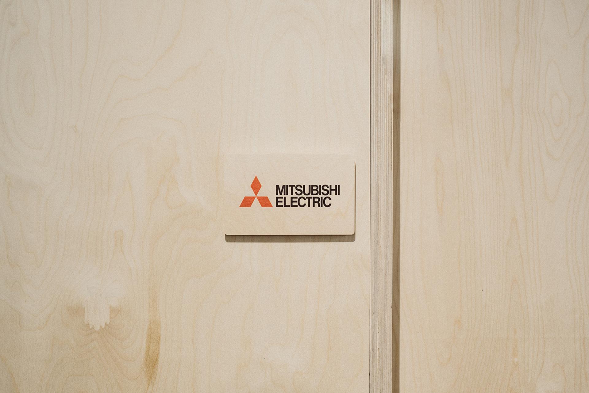 A beige birch wooden wall with a rectangular sign displaying the red Mitsubishi Electric logo and black text that reads Mitsubishi Electric, showcasing a sustainable and modern design.