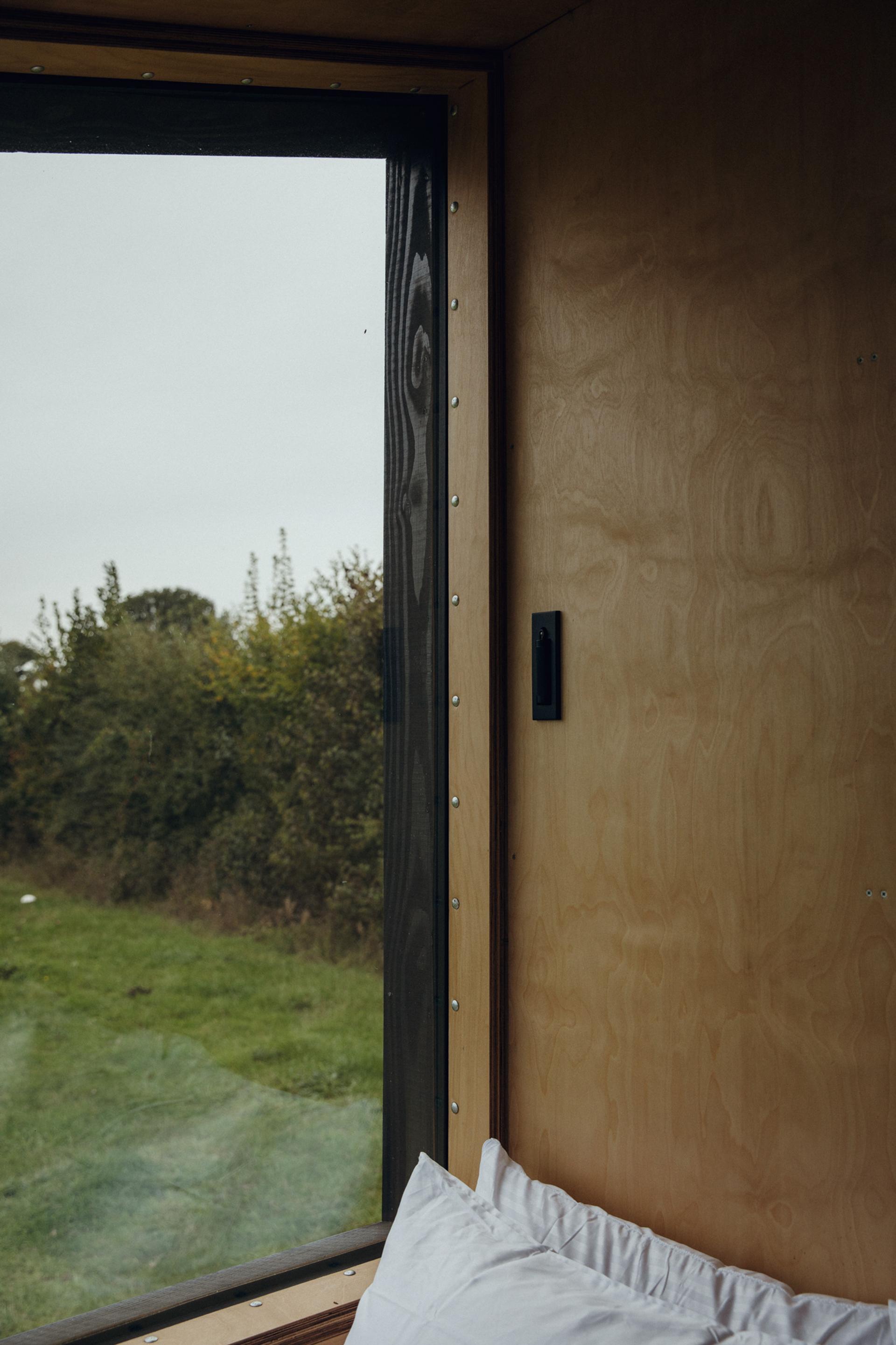 A large birch-framed window in a timber construction room overlooks a grassy field and trees; a pillow rests on a bed beneath the window, and the sky appears overcast.