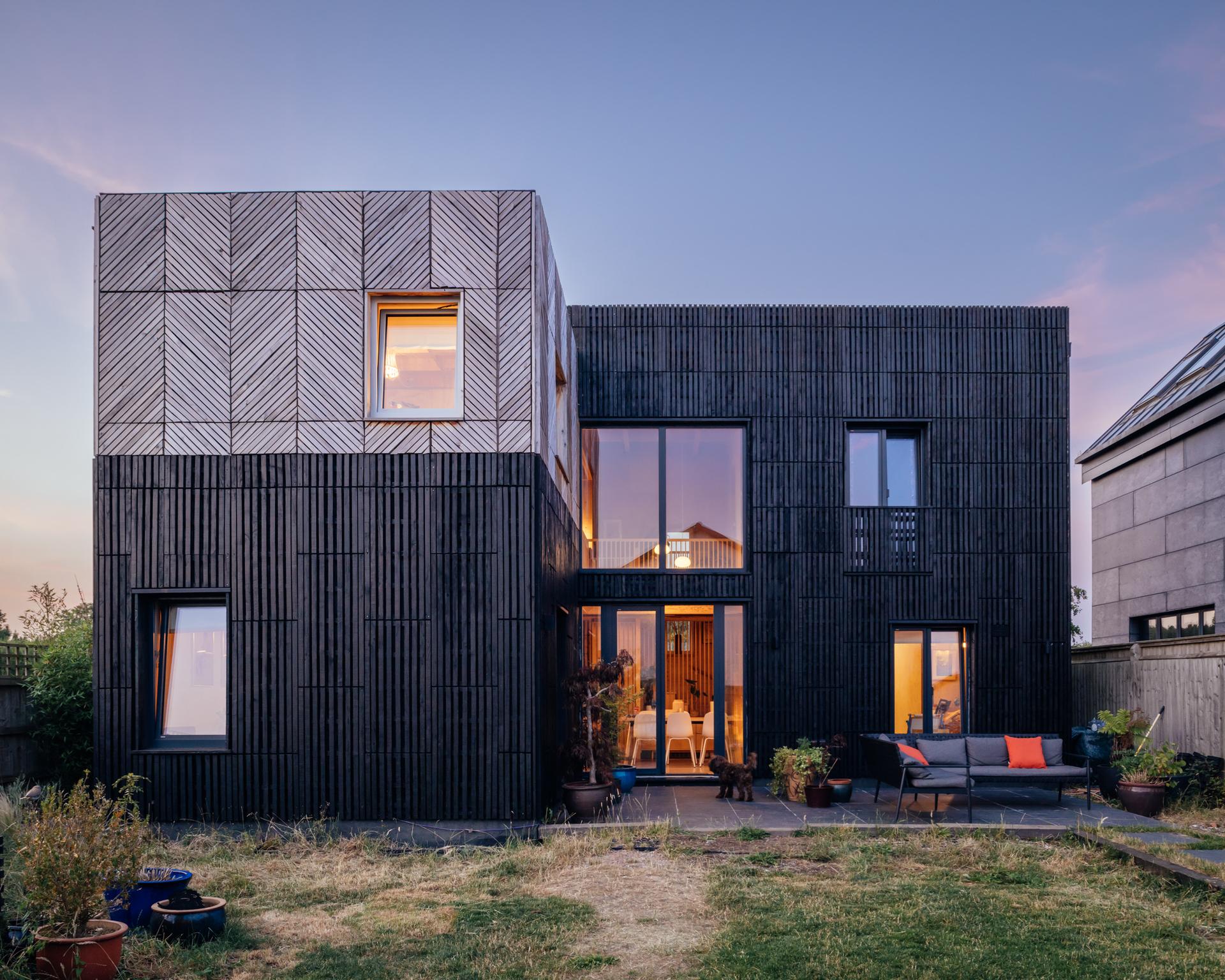 A two-storey modern house with dark vertical timber cladding and a box house extension, featuring a square upper section with chevron-patterned timber panelling, large windows, and a rear patio with outdoor seating.