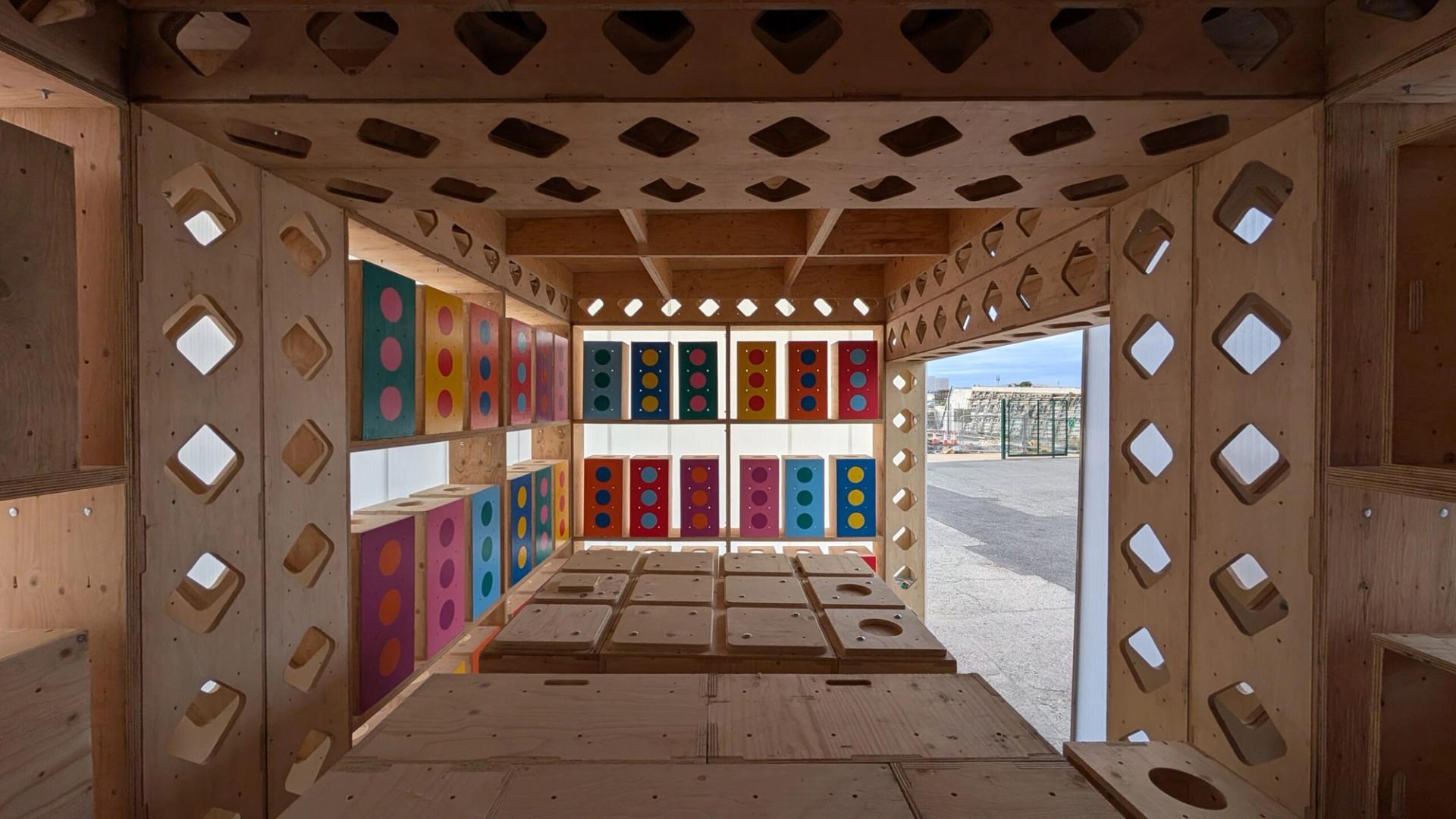 View from inside a U-Build wooden structure with square and diamond cut-outs, colourful panels with circular patterns on the walls, and sunlight streaming through openings, revealing an outdoor area beyond.