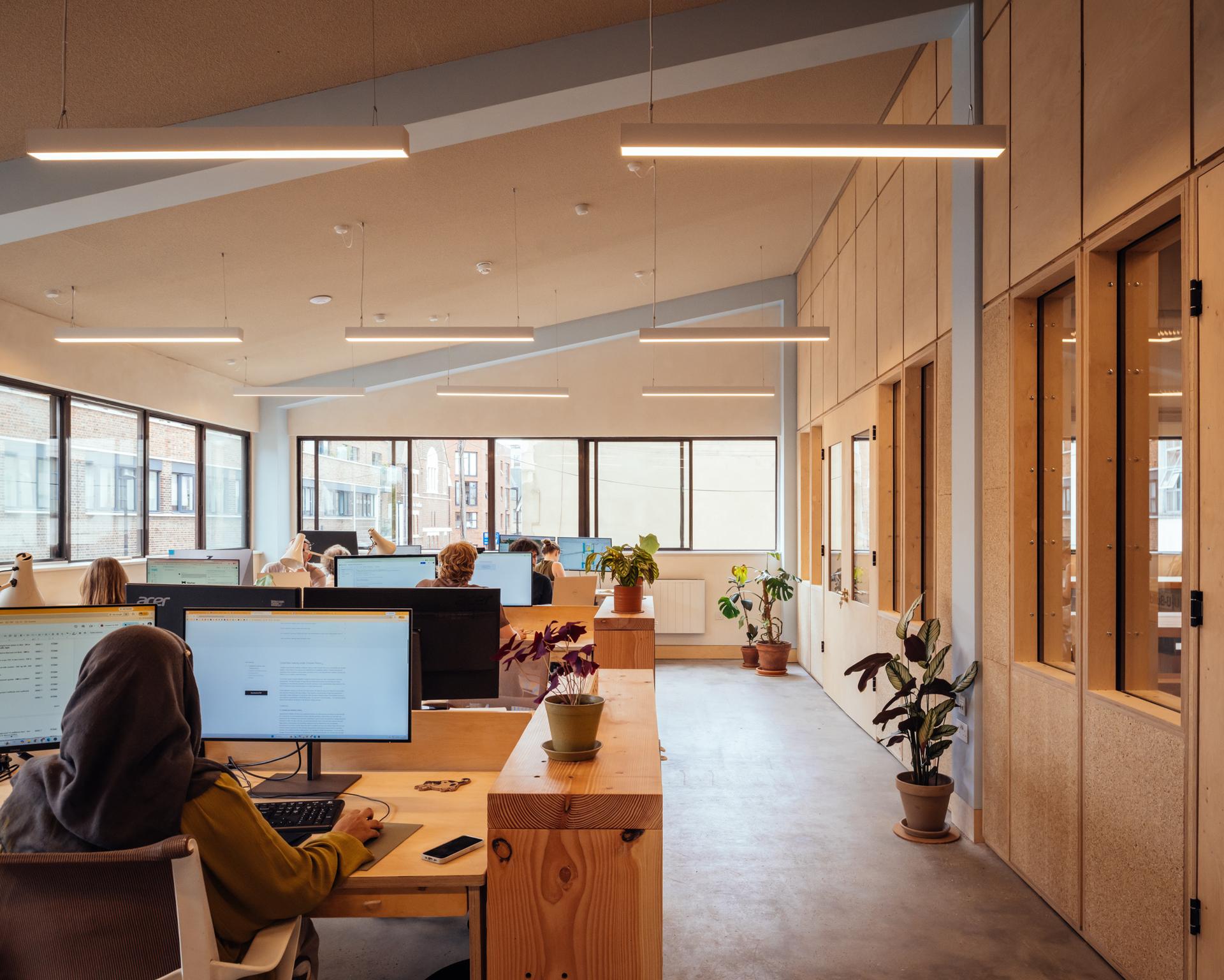 Modern open-plan office at Vyner Studios with people working at desks, large windows letting in natural light, computers, potted plants, and wooden decor elements, creating a bright and collaborative workspace.