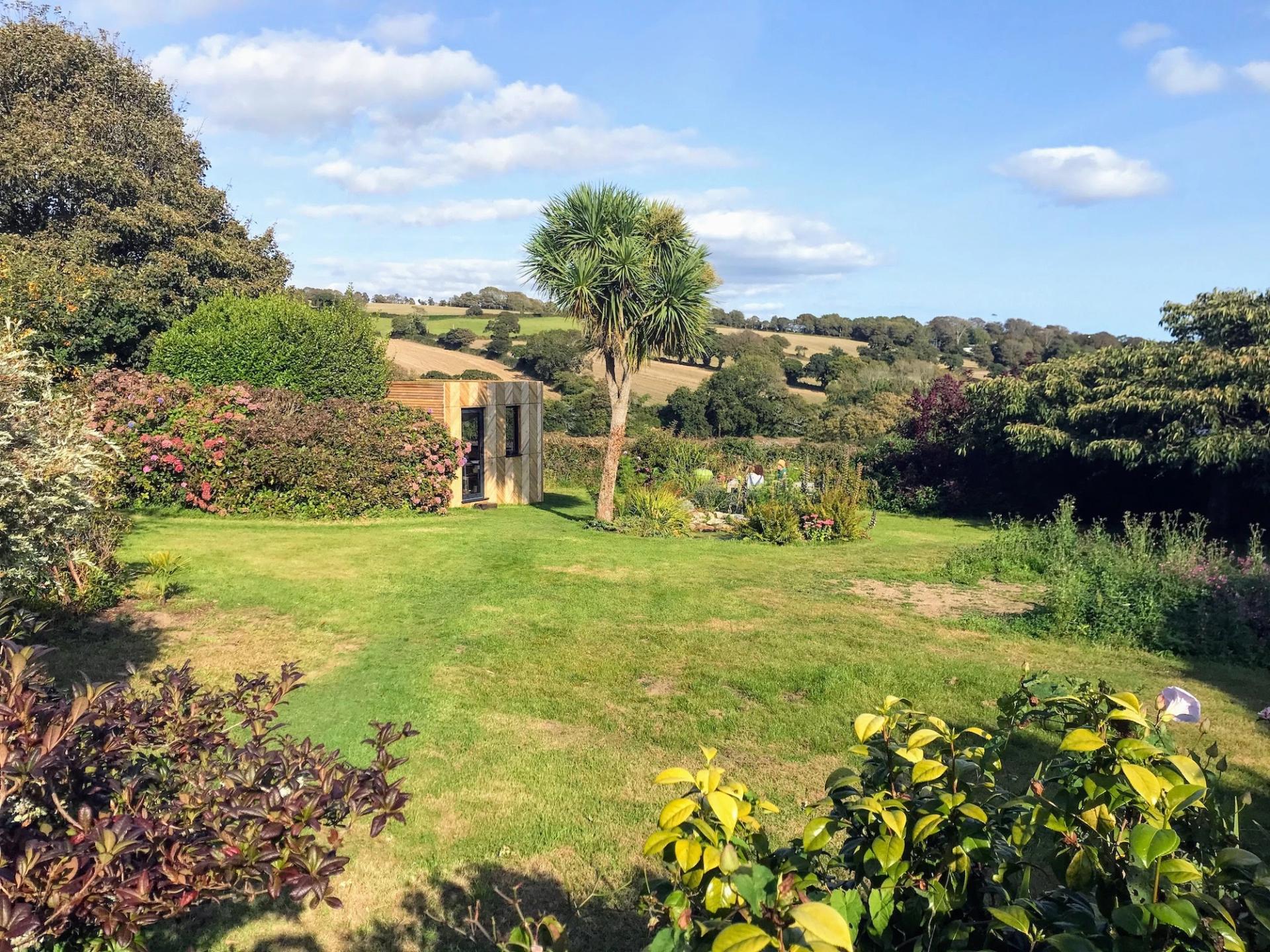 A lush garden with a palm tree, spruce, green grass, colourful shrubs, and a small wooden shed, overlooking rolling hills under a blue sky with scattered clouds.