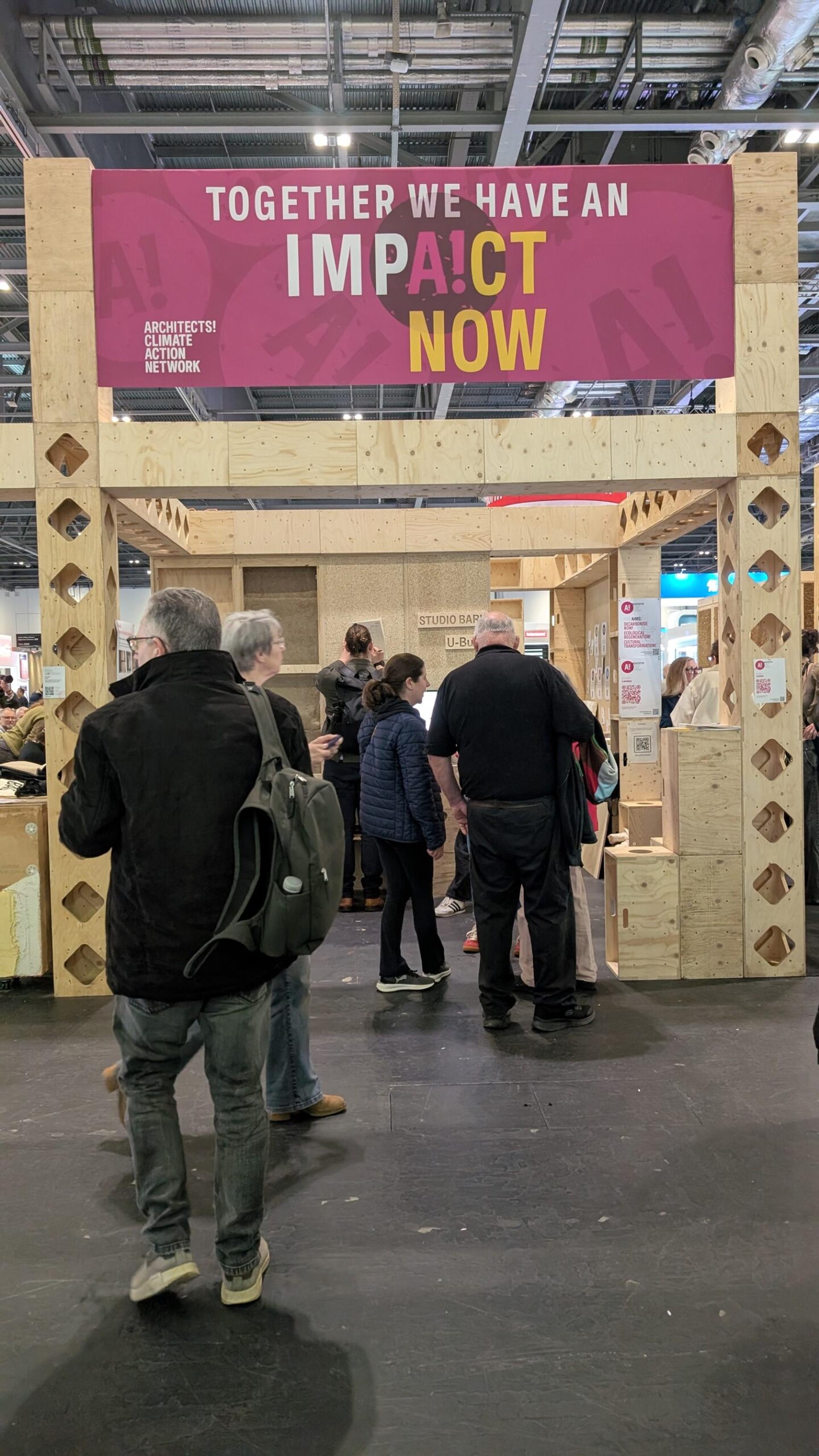 People walk through a birch plywood exhibition structure with a bright pink and yellow sign above reading, “TOGETHER WE HAVE AN IMPACT NOW,” at a busy indoor event promoting sustainable design.