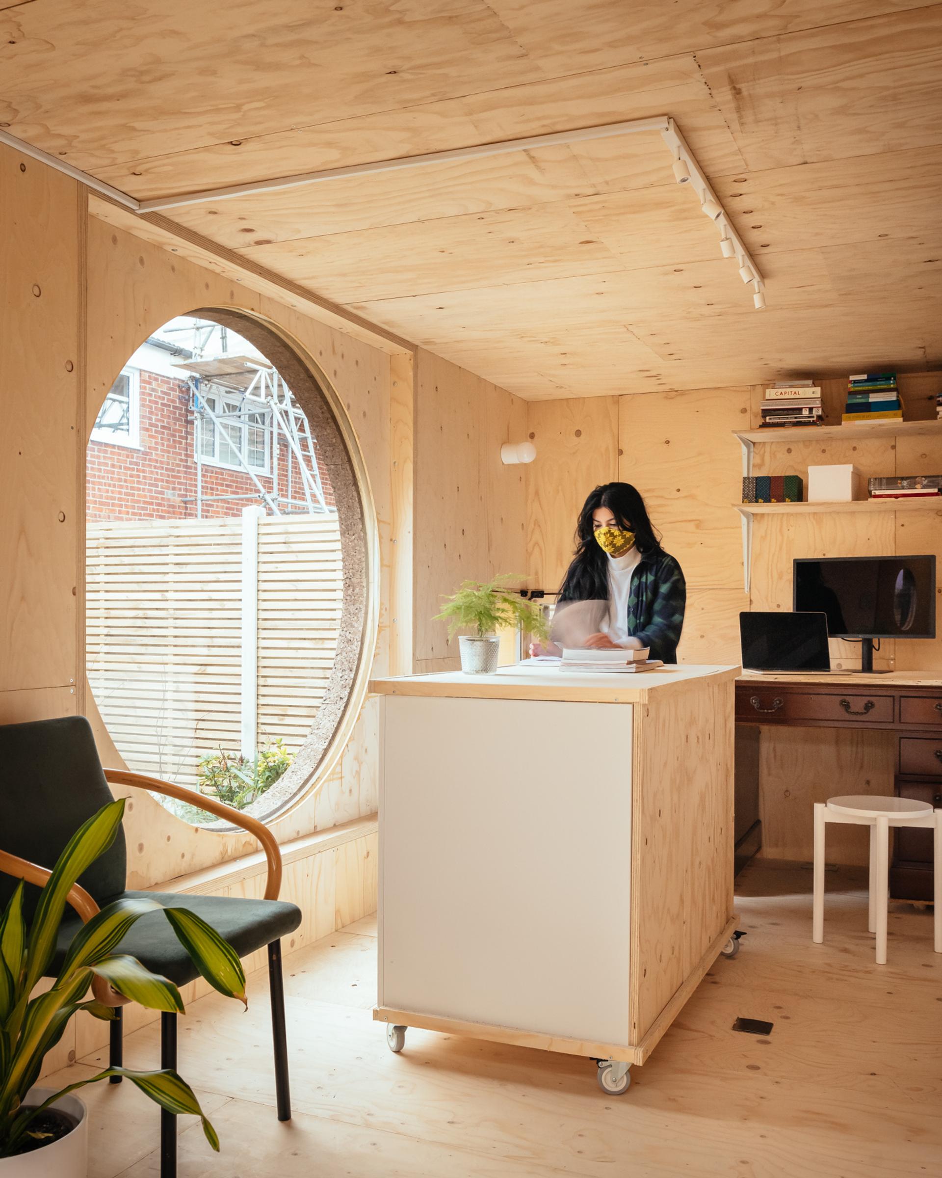 A woman stands at a plywood desk in a modern, minimalist office with light wood walls, a round window, and various plants and shelves, creating a cosy and bright workspace.