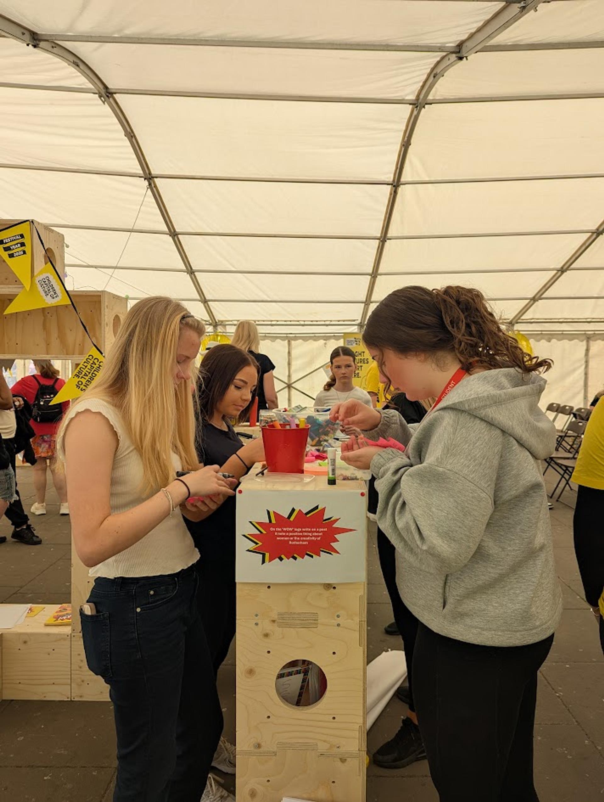 Three young women stand at a wooden table in a marquee, focused on crafts or writing activities. The table, made from sturdy birch plywood, features a colourful sign on the front. Other people and chairs are visible in the background.