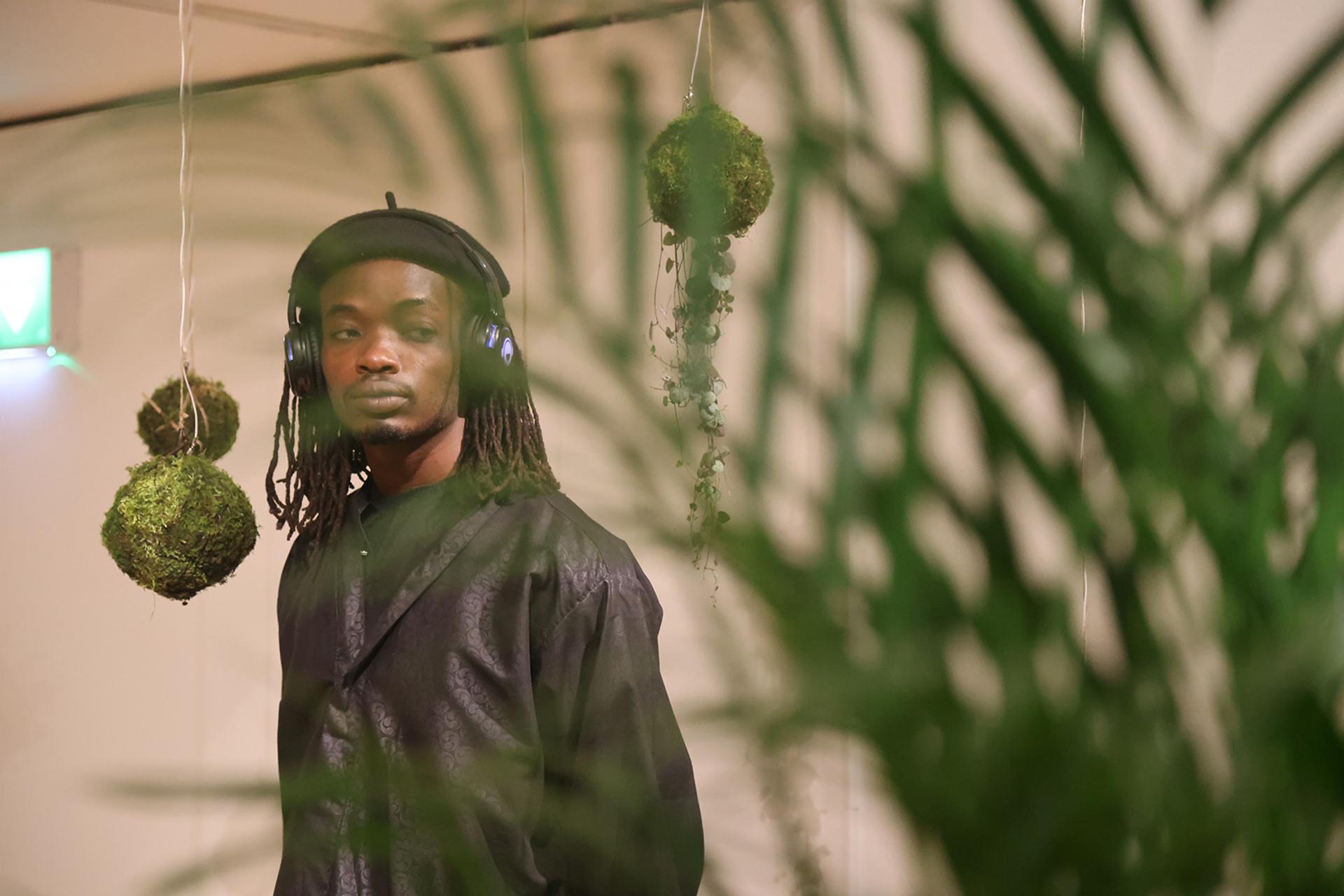A person wearing headphones and a dark outfit stands indoors, partially obscured by green leafy plants. Moss balls hang from the ceiling in the background, creating a sustainable, serene atmosphere inspired by spruce and birch forests.