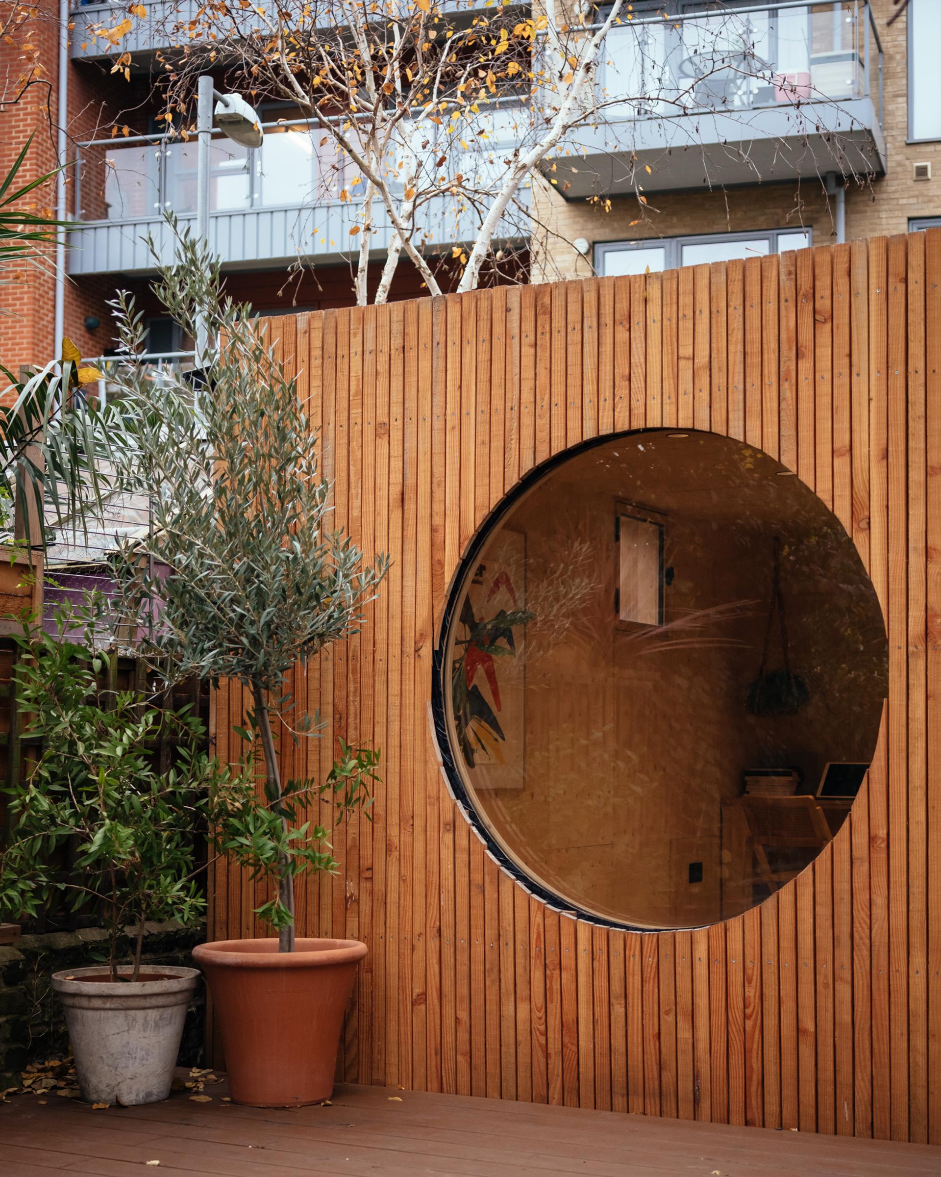 A modern birch plywood structure with vertical slats features a large circular window. In front, a potted tree sits on the wooden decking, while contemporary block of flats can be seen in the background.