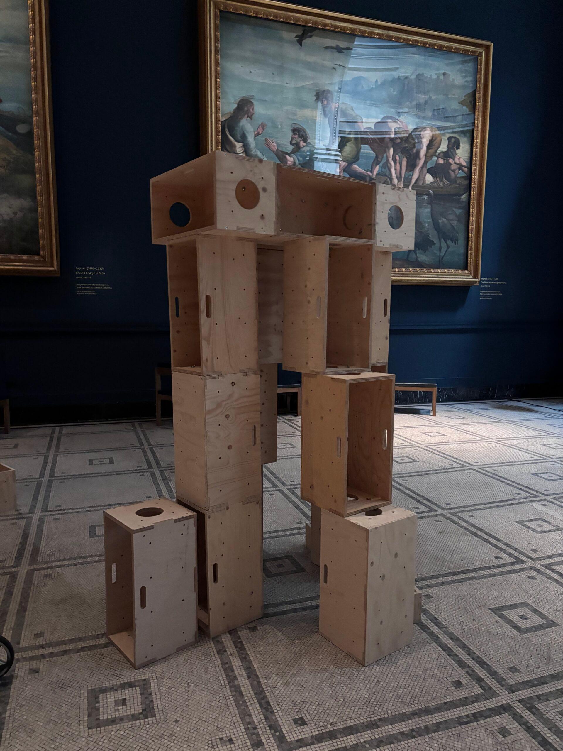 A sculpture made of stacked wooden crates stands in a gallery at the V&A, surrounded by classical paintings on the blue walls and a patterned tiled floor—a striking example of how upstart artists build structures for activism within traditional spaces.