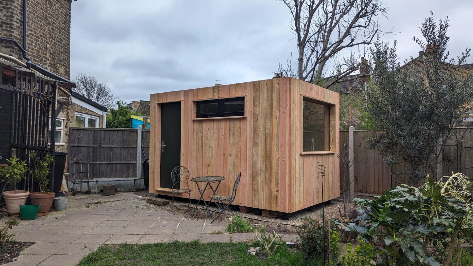 A small modular wooden shed with large windows and a door stands in a garden near potted plants and outdoor furniture, surrounded by a fence and neighbouring houses.
