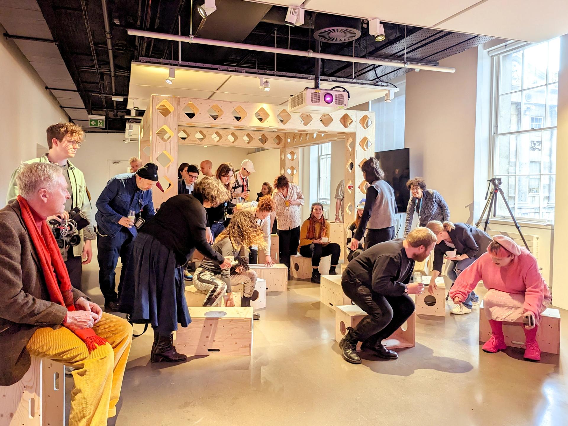 A group of people interact and converse in a bright, modern room with modular wooden box seating and geometric decor. Some are standing or bending over, whilst others are seated, all engaged in a lively, social atmosphere.