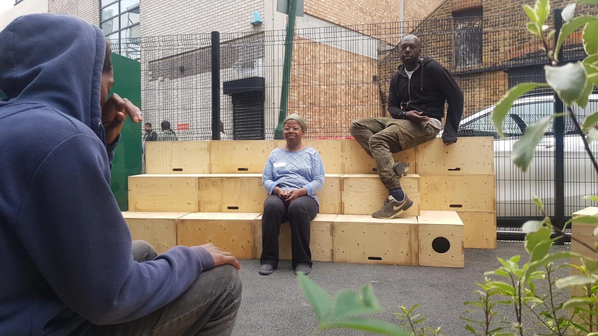 Three people sit and talk on wooden steps outside, near a fence and brick building. One woman sits in the centre, while two men—one in a hoodie, the other in a jacket—chat about ways to recycle and upcycle. Plants grow in the foreground.
