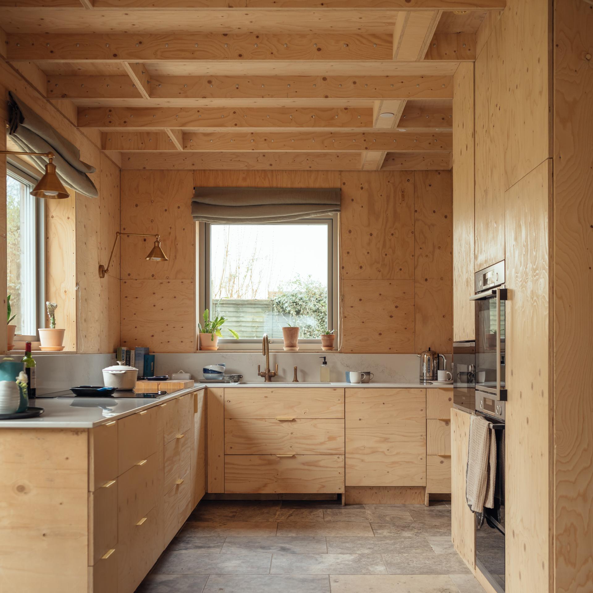 Modern kitchen with light plywood cabinetry and exposed timber beams, featuring a sink, worktops, and a wall-mounted oven.