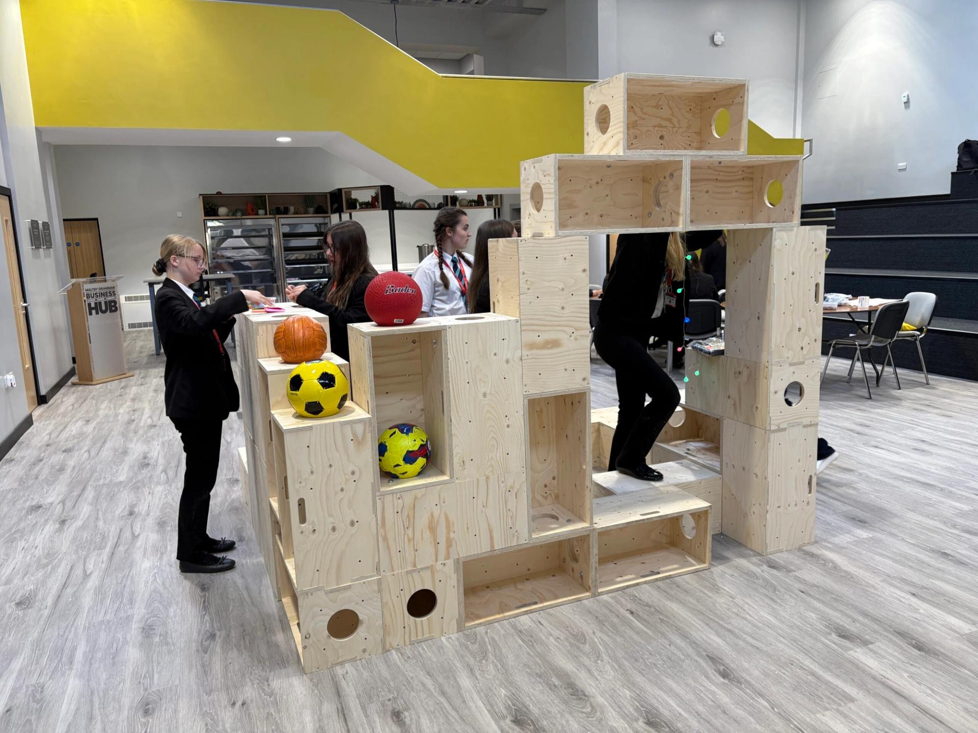 Students in uniform interact around a large modular birch play structure with various colourful balls and objects placed in its cubby holes; some students are writing or reading near the sustainable structure in a modern, bright room.