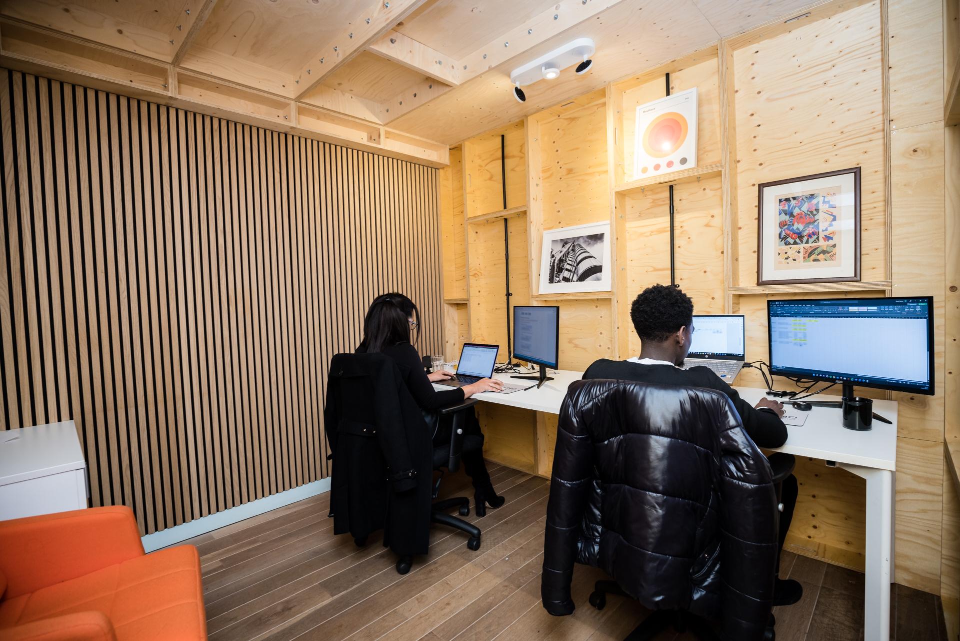 Two people work at separate desks with computers in a small, modern office featuring birch plywood walls, framed artwork, and overhead lighting. An orange chair is in the foreground.