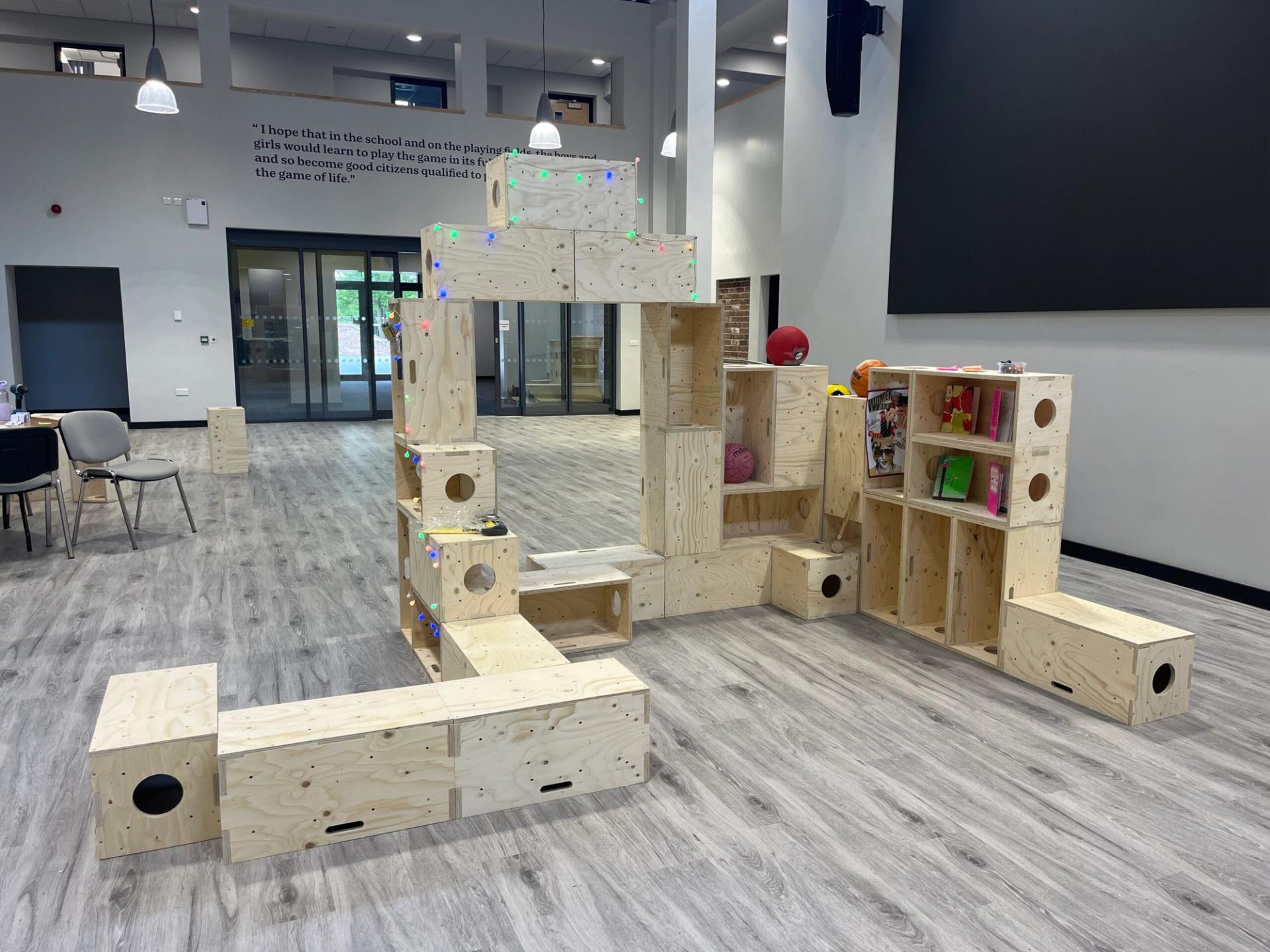 Large indoor playroom featuring stacked, modular birch wooden blocks arranged as a play structure with holes and coloured lights. Modern decor, grey wood flooring, chairs, and an inspiring quotation on the wall in the background.