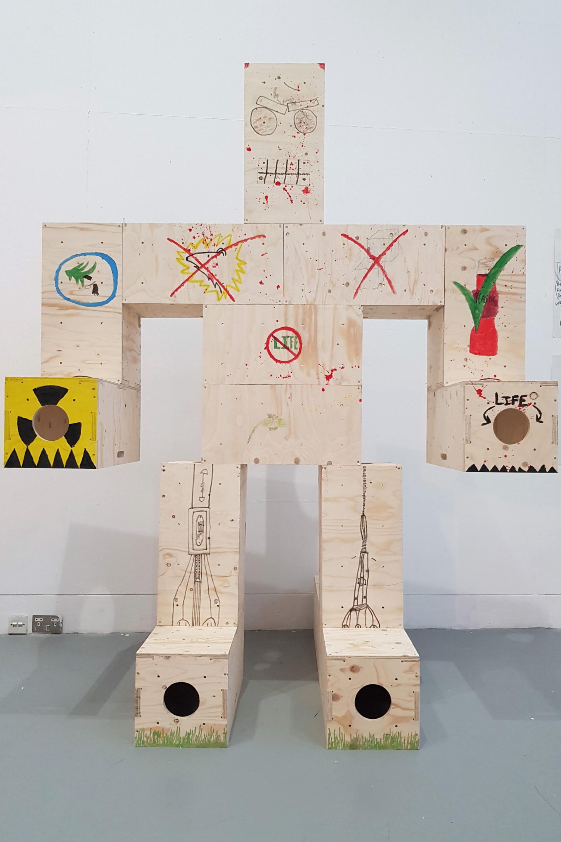 A large U-Build humanoid figure made of stacked wooden boxes, each decorated with faces, plants, symbols, and abstract markings, stands against a plain white wall.