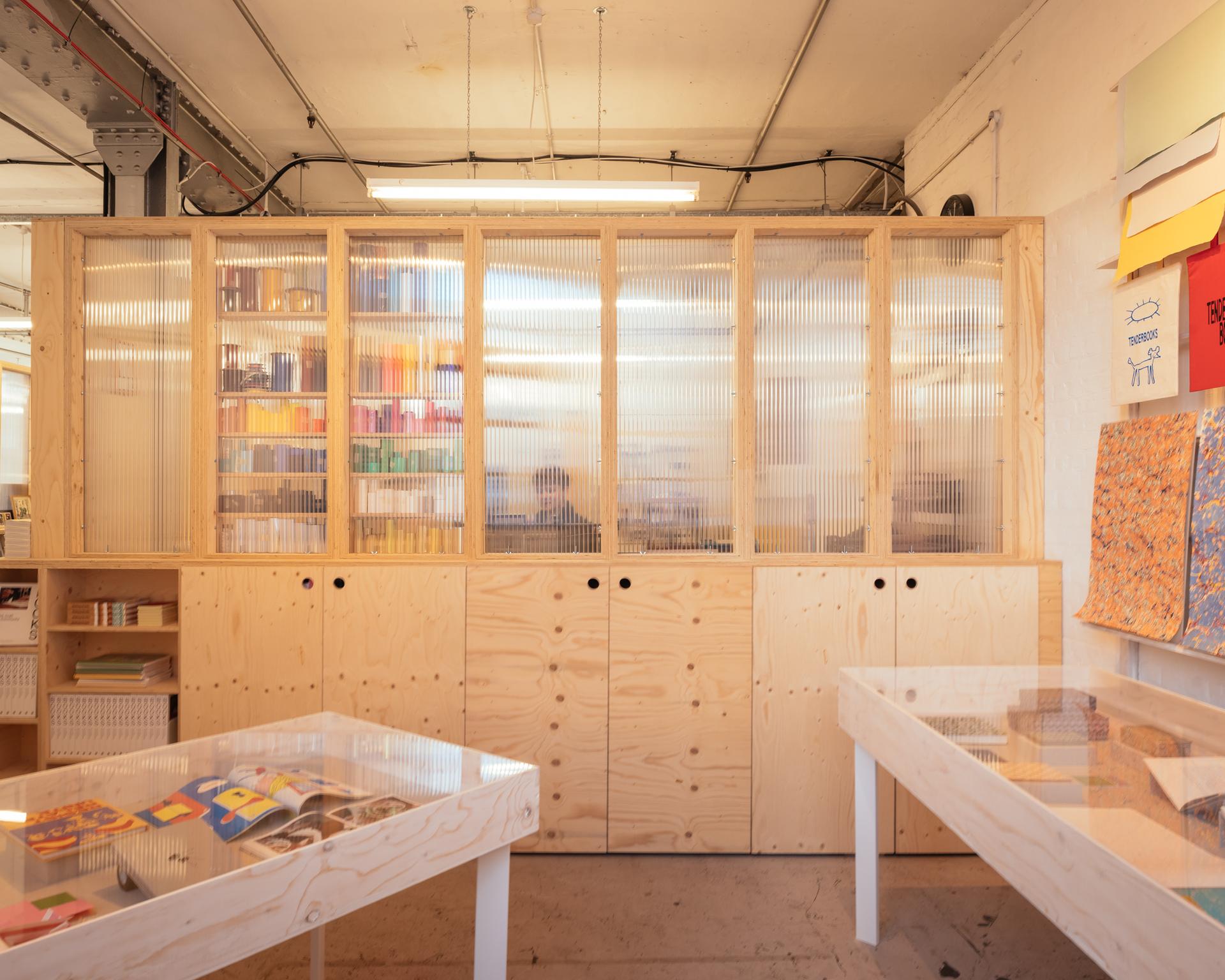 A bright, organised workspace with modular wooden cupboards and translucent panels, displaying colourful papers and books. Two tables with clear tops hold various art supplies and creative materials, all highlighting sustainable timber construction.