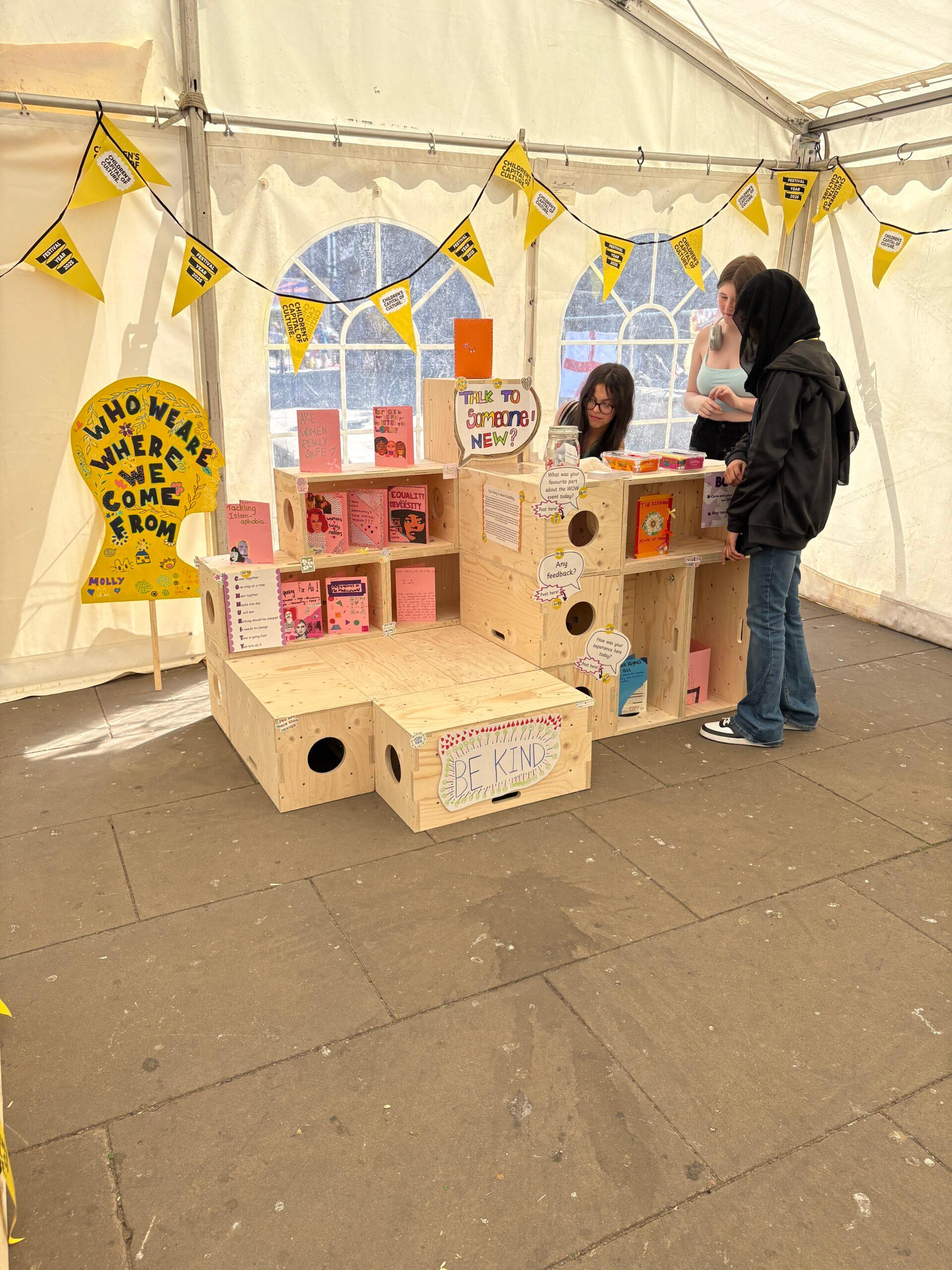 Three people are at a modular plywood stall inside a marquee decorated with yellow bunting. The stall displays colourful books and handmade signs, including “BE KIND” and “WHO ARE WE? WHERE HAVE WE COME FROM?”.