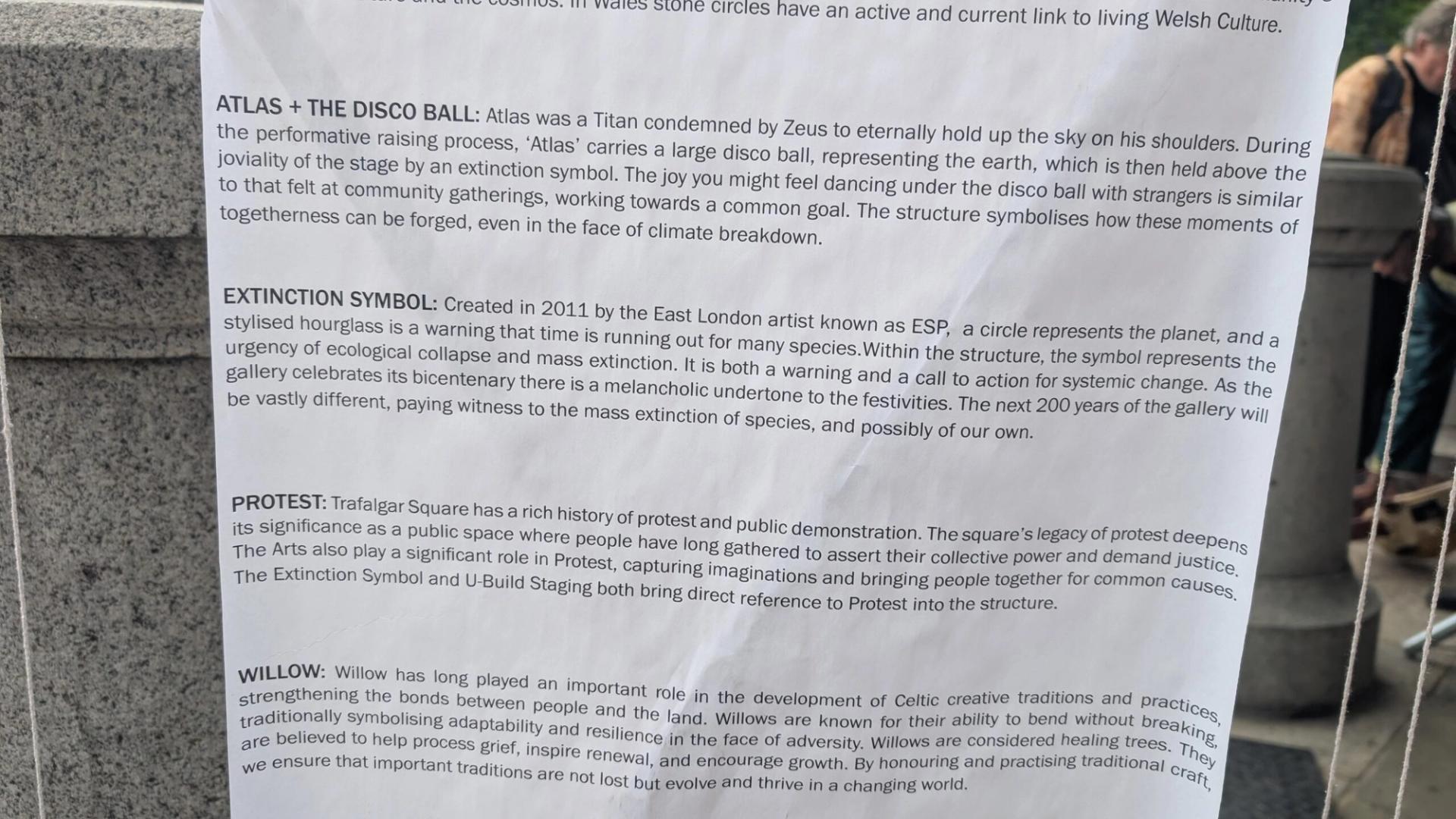 A close-up of an informational sign describing four art installations—Atlas + The Disco Ball, Extinction Symbol, Protest, and Willow—featured in the 2025 Triumph of Art event at the National Gallery, attached to a stone surface outdoors.