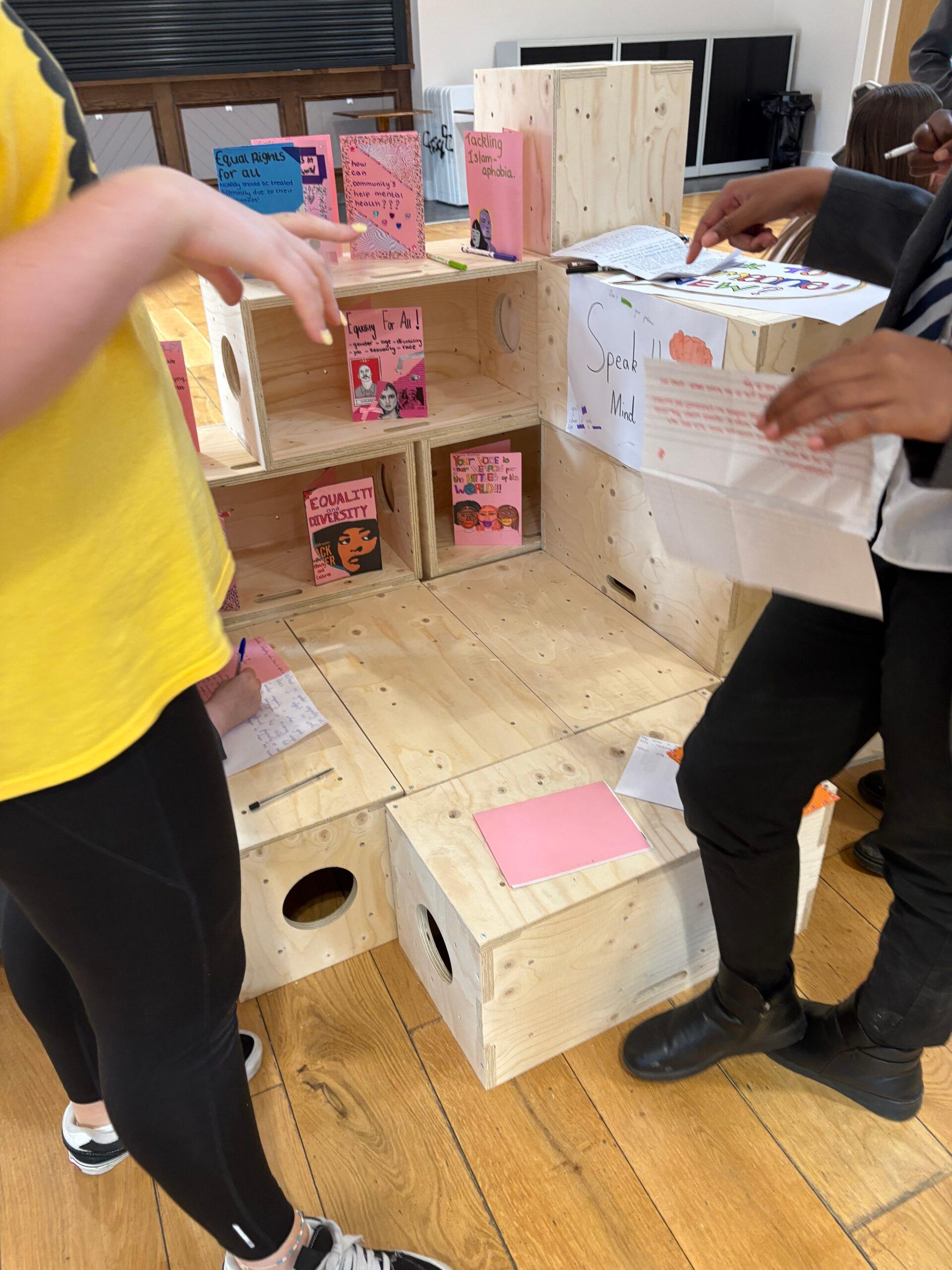 Three people stand near a modular wooden bookcase display filled with colourful books and papers. Two hold papers, and one wears a yellow shirt. The timber construction of the floor adds to the sustainable feel, with computers visible in the background.