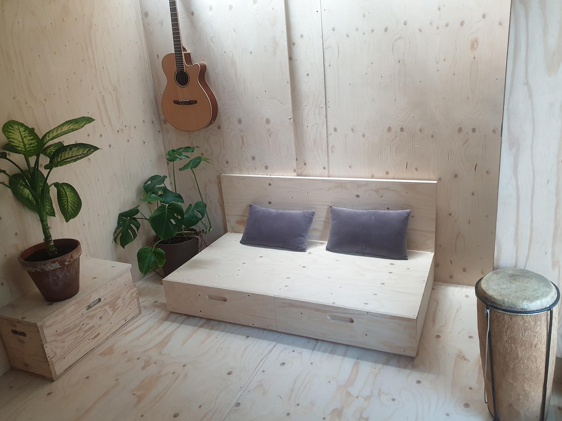 A minimalist wooden room features a small sofa with two grey cushions, potted plants, a drum, a wooden box made from recycled materials, and an acoustic guitar mounted on the wall—showcasing subtle touches of sustainability.