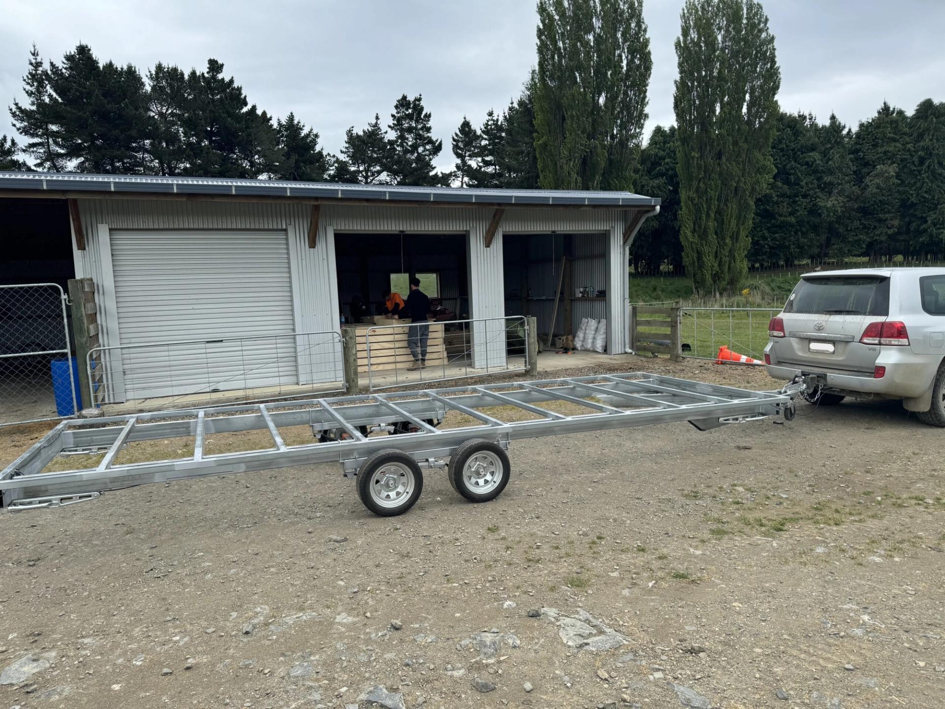 A silver trailer chassis is attached to a white SUV parked on a gravel drive outside a metal shed. Several people are working near the shed entrance, which is surrounded by trees and lush greenery, highlighting a sustainable timber construction project.