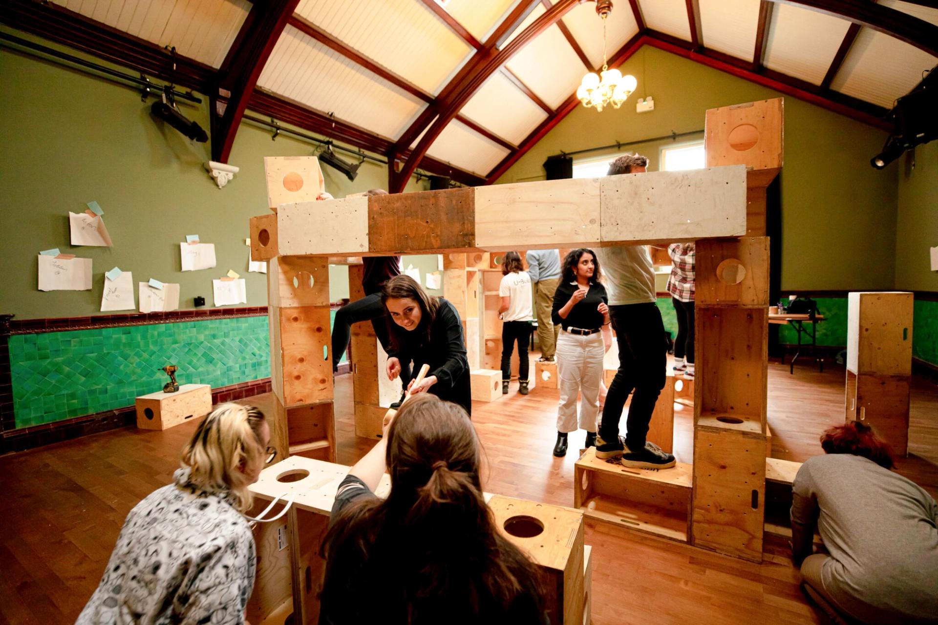 A group of people collaborate to build a large modular structure from spruce wooden blocks in a spacious, high-ceilinged room with green walls and wooden floors. Some are standing on blocks whilst others are assembling pieces.