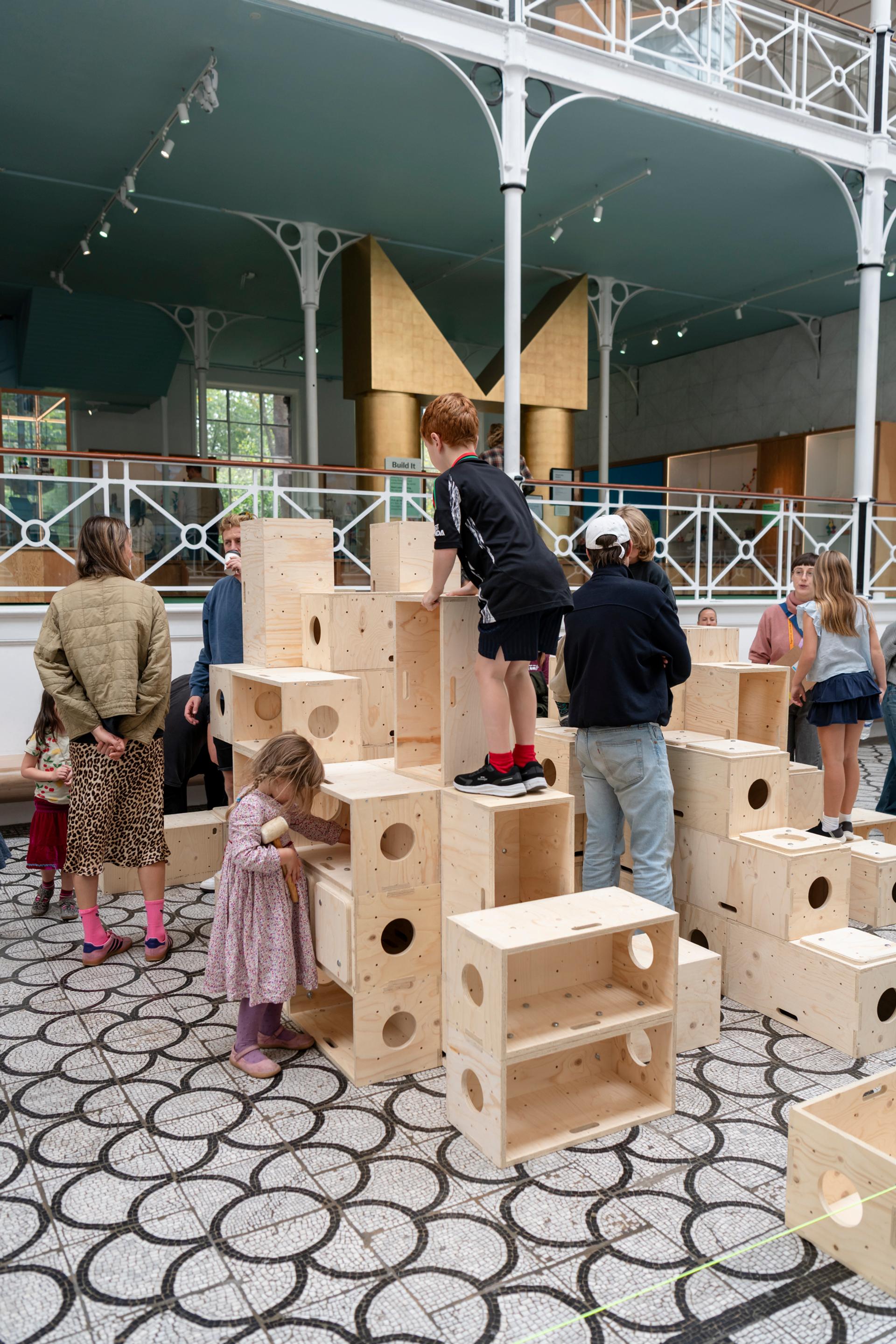 Children play and climb on large U-Build wooden blocks in a spacious indoor area with patterned flooring, surrounded by adults and other children. The setting appears to be an interactive exhibition or play space.