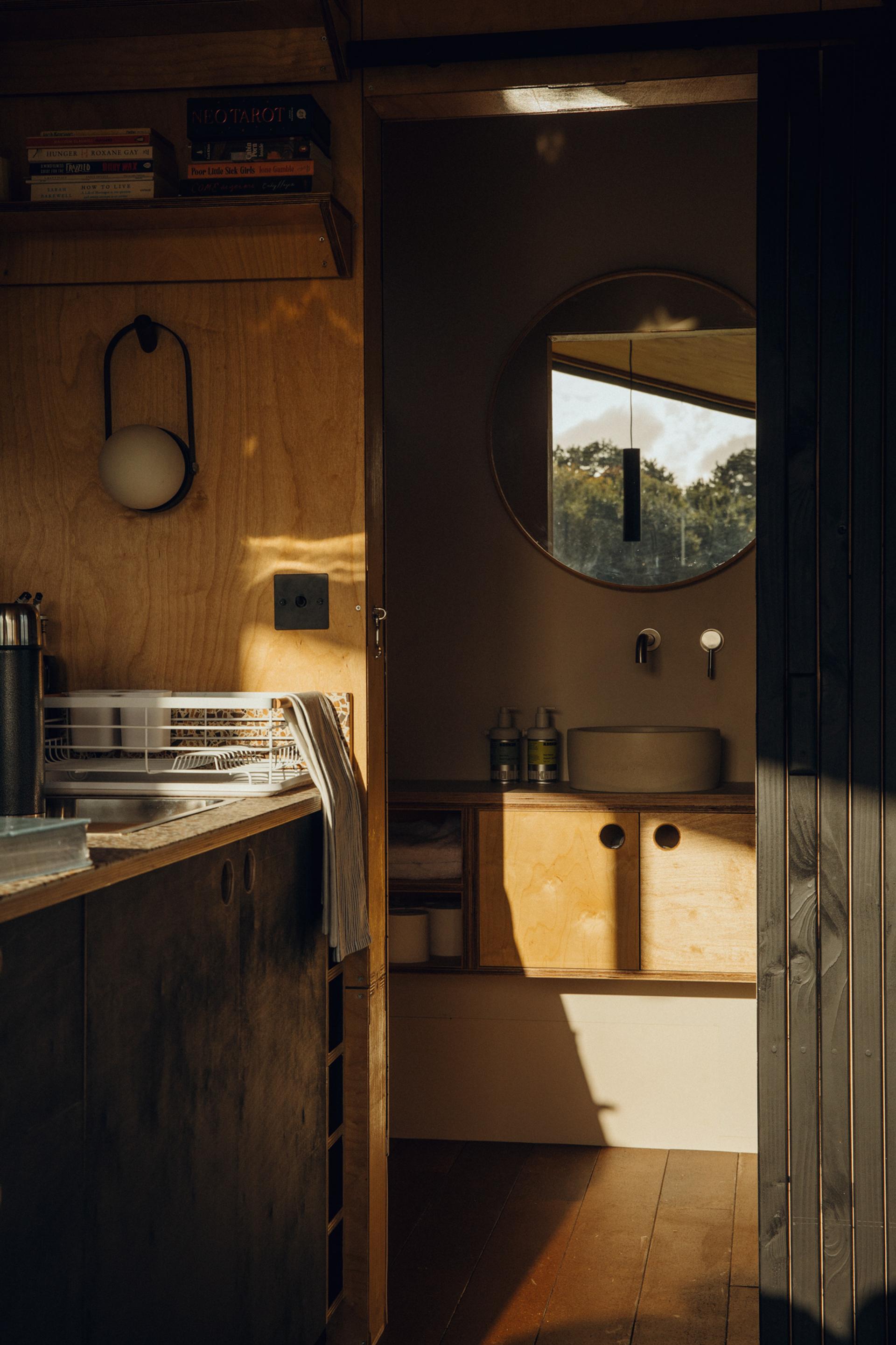 A cosy, sunlit bathroom with birch wooden walls and cupboards, a round mirror above a small basin, shelves with towels and bottles, and part of a sustainable kitchen with books and a dish rack visible in the foreground.