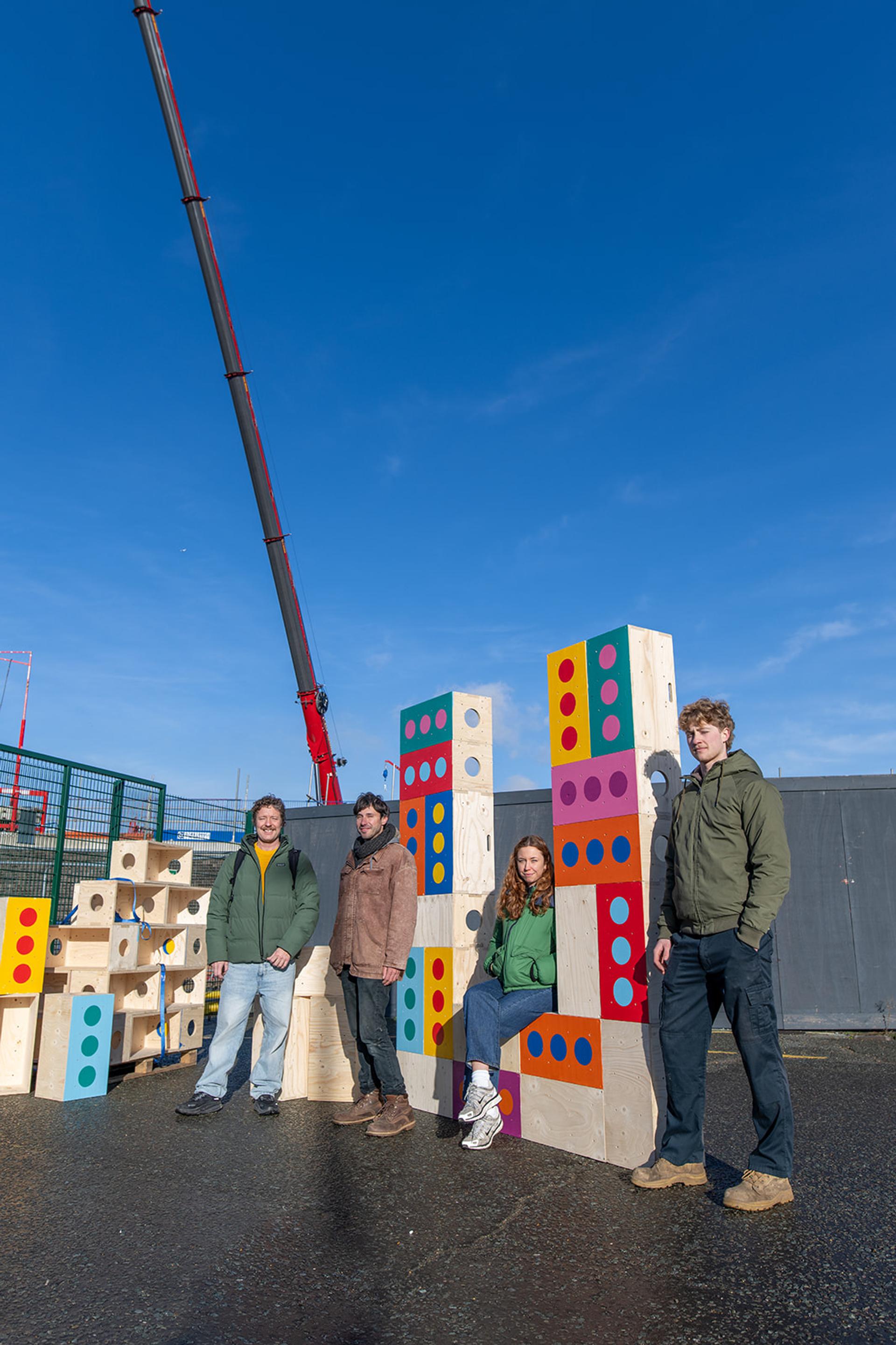 Four people in winter clothing stand outdoors beside large, colourful wooden blocks stacked to resemble dominoes, capturing a playful scene. A tall red crane stretches into the blue sky, near what appears to be a community Box Library.