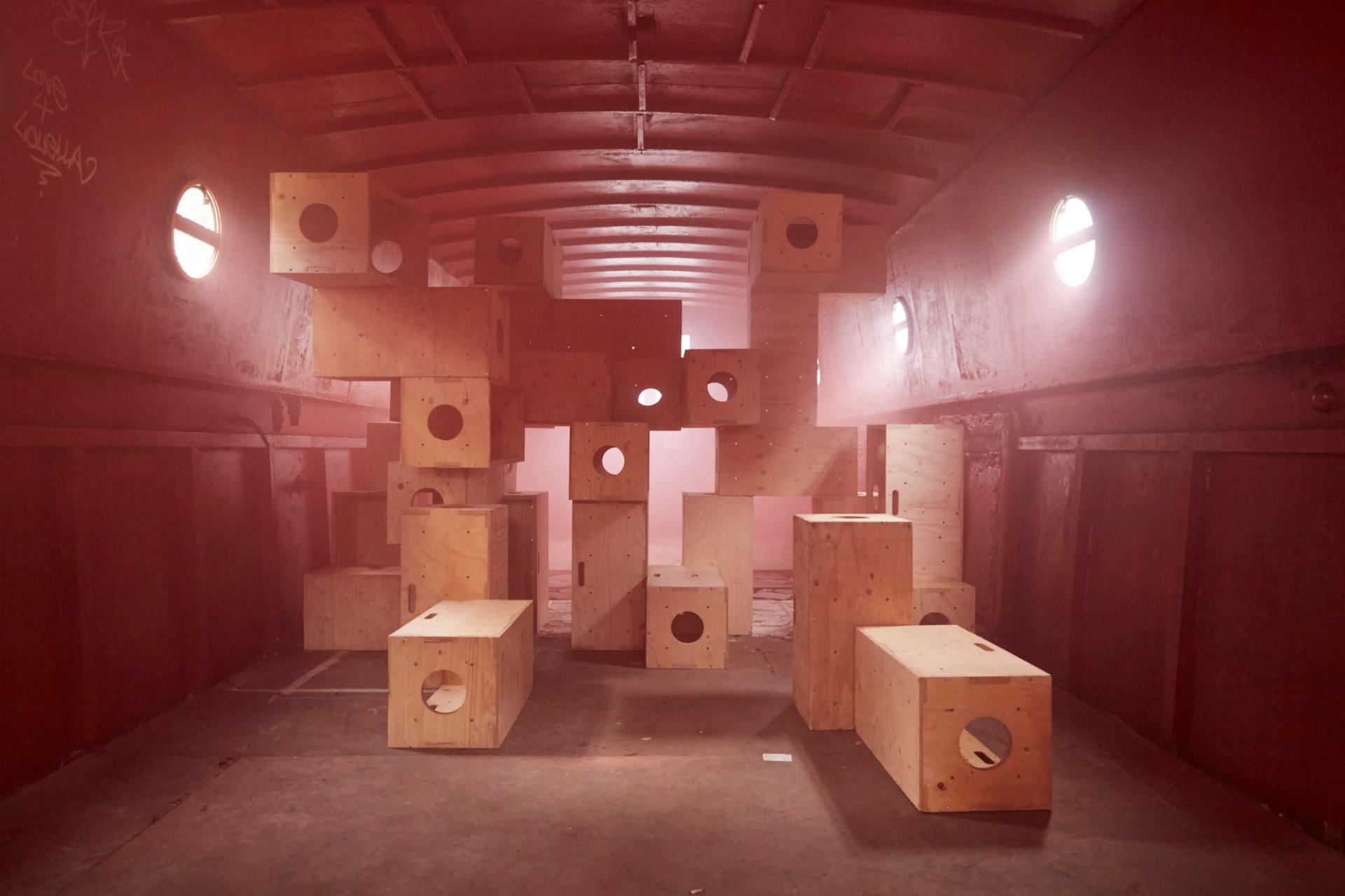 The central stack of plywood modules, now captured in a soft, hazy atmosphere. The lighting is diffused, pink and warm, giving the scene a dreamy, monochromatic red and gold quality. The circular cutouts in the boxes align to create a sense of depth through the center of the stack.