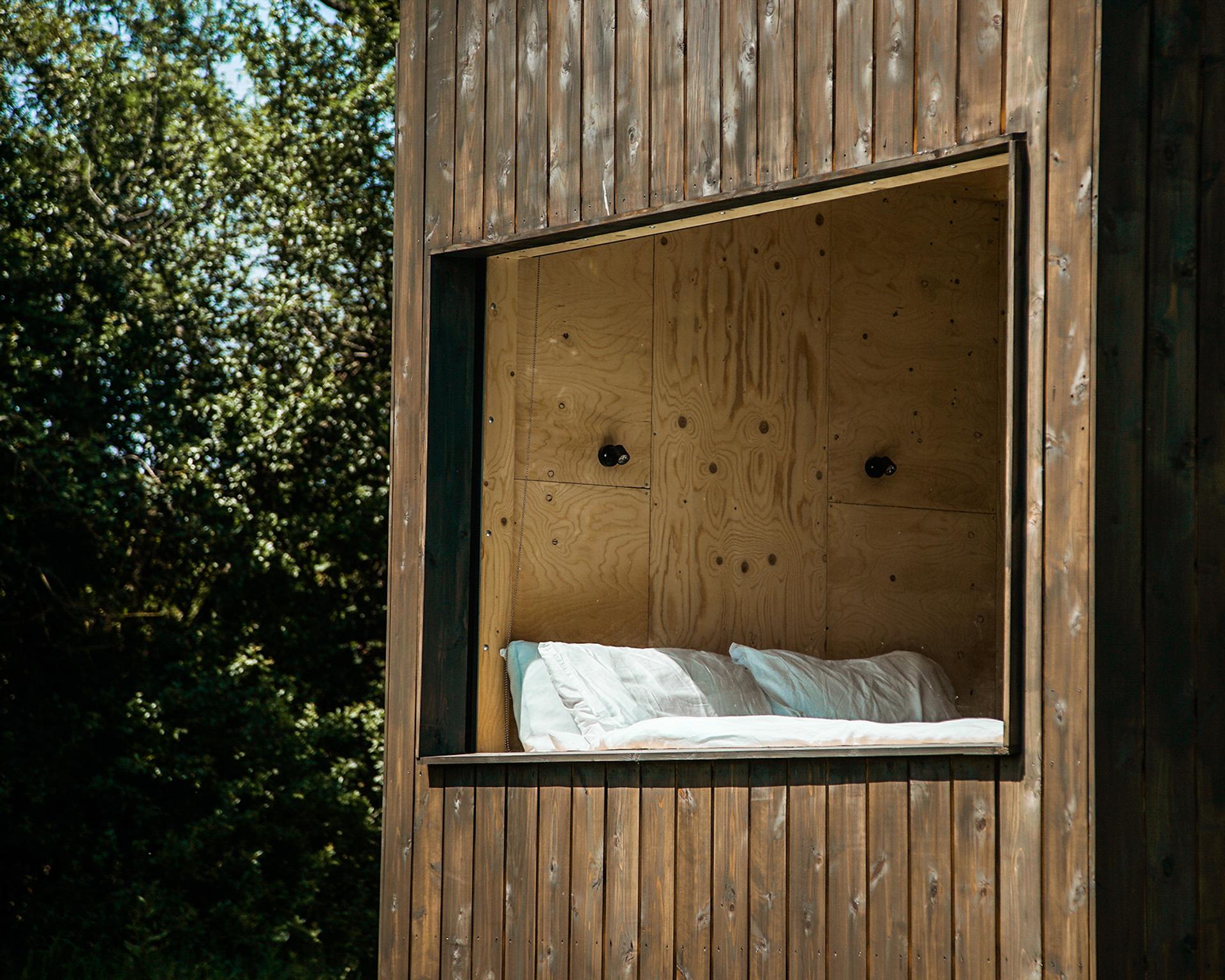 A cosy outdoor nook with white pillows and bedding is built into a modern wooden structure using sustainably sourced materials, with a large square window opening to trees and greenery outside.
