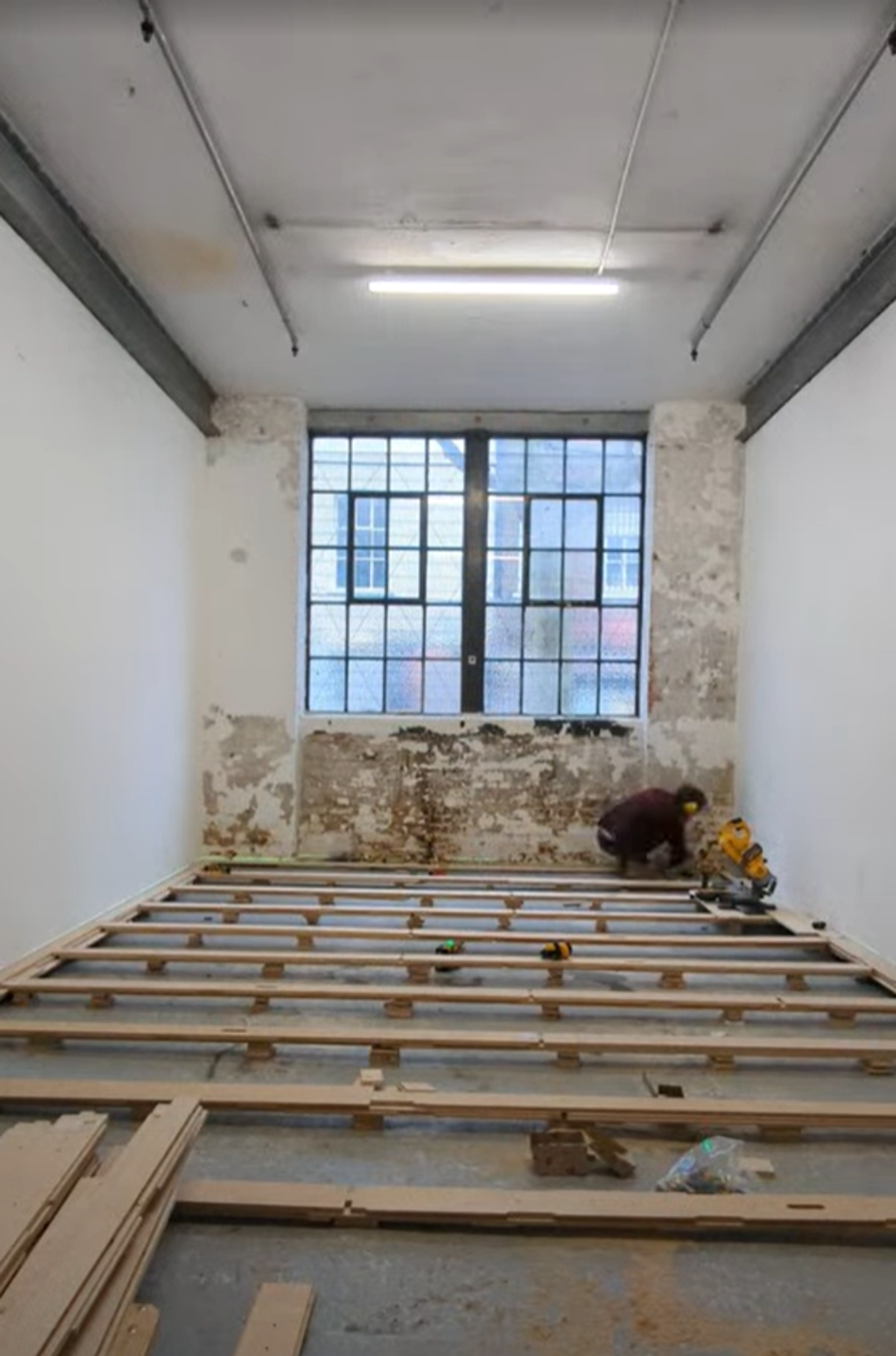 A person kneels on the floor in a long, narrow room with exposed beams and partially stripped walls whilst building a modular wooden framework. Large industrial windows let in daylight.