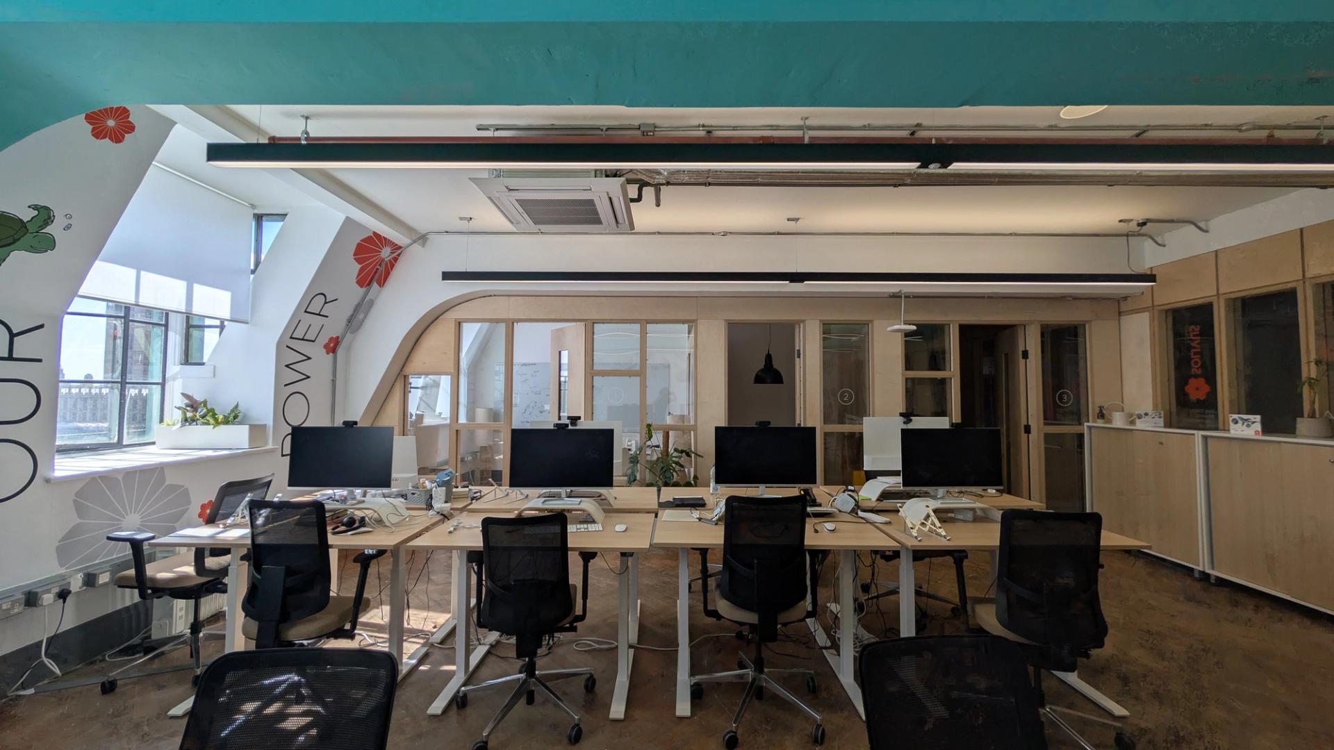 A modern, modular open-plan office with several desks, computers, and chairs, large windows allowing natural light, plants, and light birch cupboards along the back wall. The atmosphere is bright and organised.