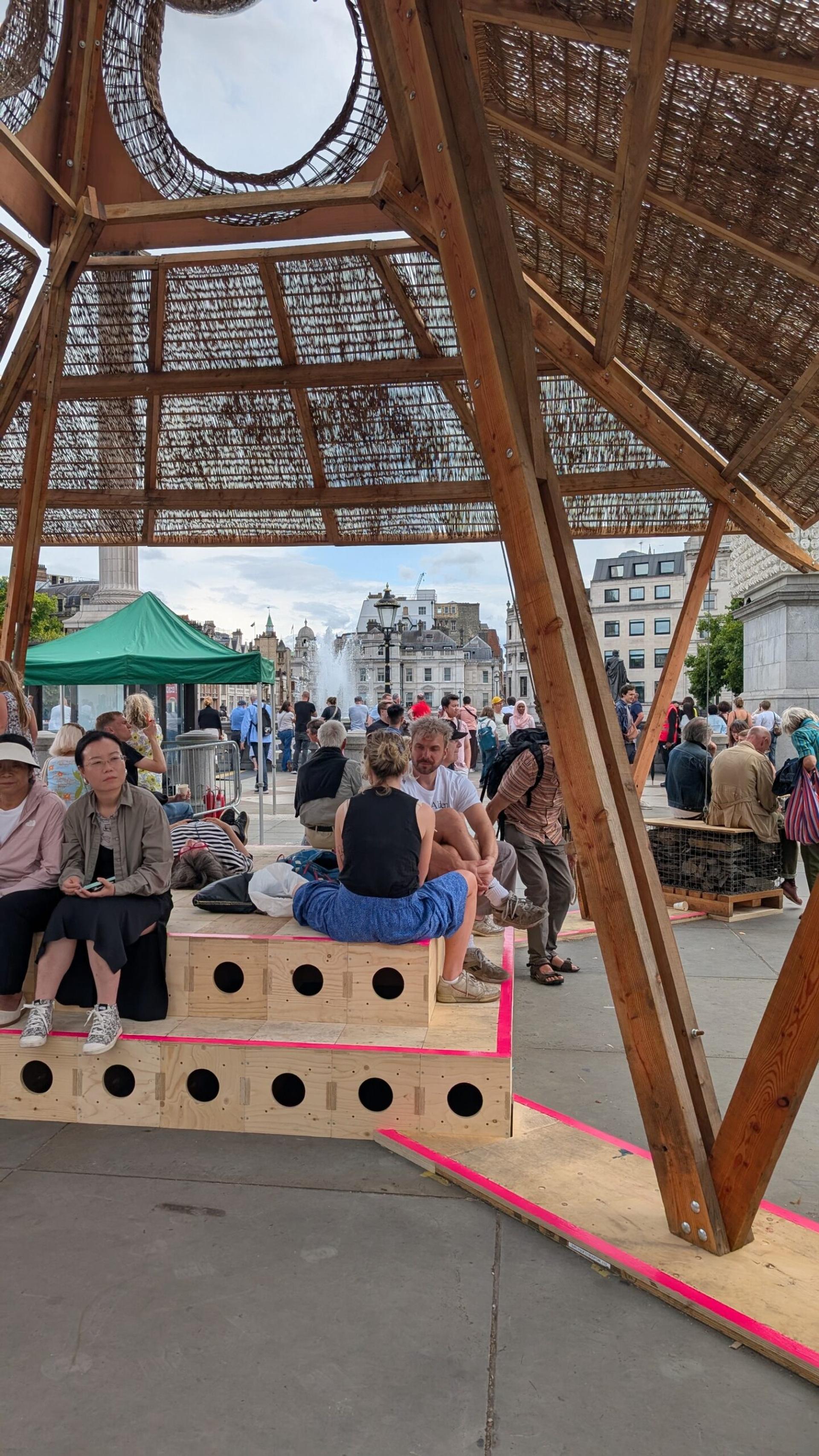 People sit and relax on wooden benches, made from upcycled materials, under a large wooden canopy in a busy outdoor public square, with a green marquee, fountain, and buildings visible in the background.