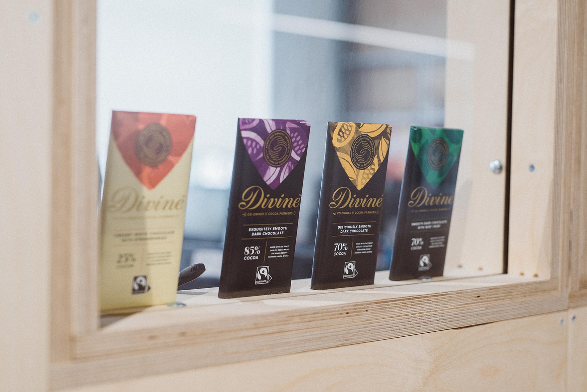 Four Divine chocolate bars in vibrant wrappers (gold, red, purple, green) stand upright on a light spruce shelf, viewed through a glass window.